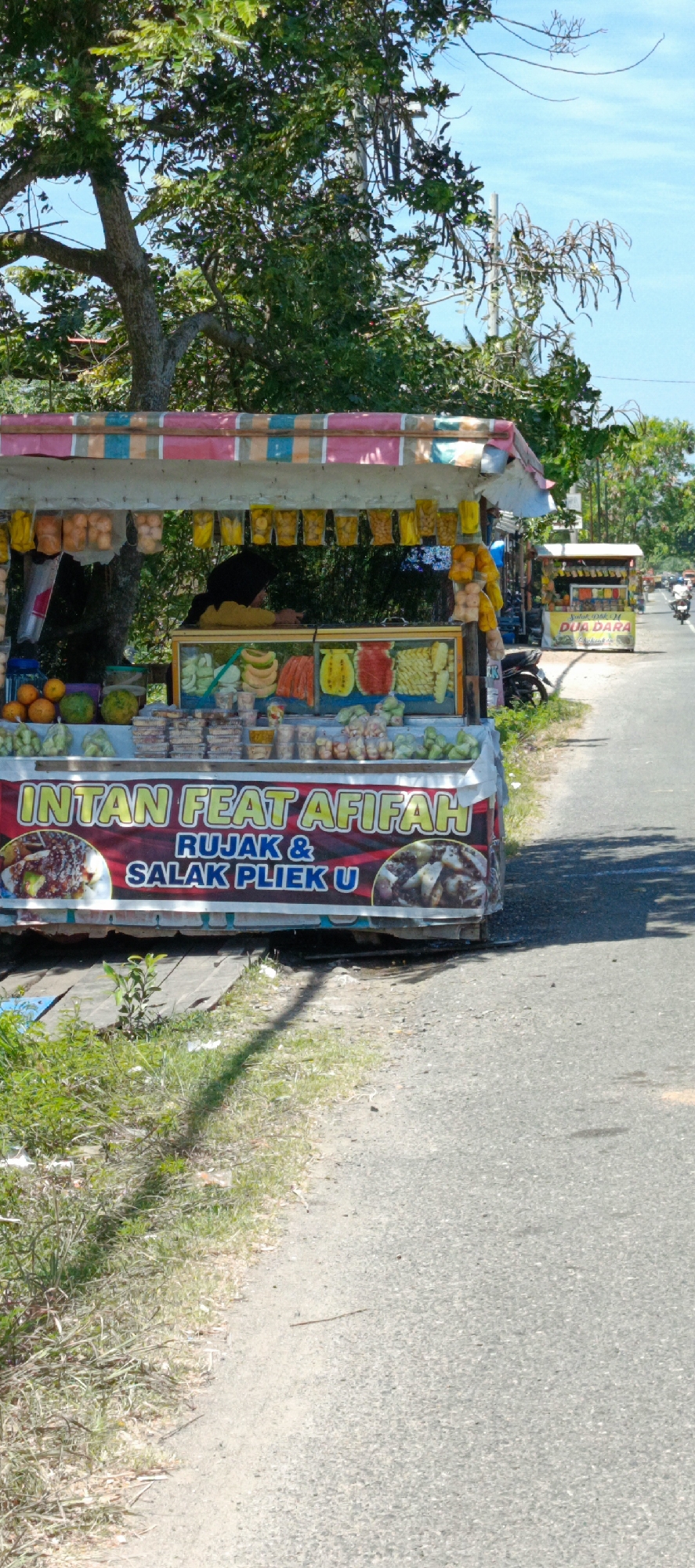 A roadside fruit stand is pictured in Lampisang, Indonesia, on a sunny day. The stand, named "INTAN FEAT AFIFAH," displays a variety of fruits and snacks. In the foreground, a colorful banner advertises "RUJAK & SALAK PLIEK U." The stand is adorned with a striped awning and various packaged goods. Behind the fruit display, a person wearing a yellow shirt and a headscarf is visible. To the right of the frame, another fruit stand is visible further down the road. The scene depicts a typical Indonesian roadside market with its vibrant offerings and bustling atmosphere.