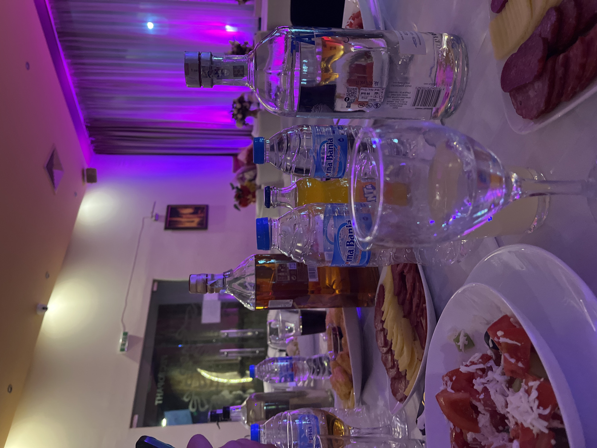 The image depicts a table setting, likely at a restaurant or event, in an unidentified location that may resemble a scene in Elhovo, Bulgaria. The table is covered with various beverages, including a bottle of Absolut vodka, bottles of "Voma Bania" mineral water, and a bottle of an amber-colored spirit, possibly whiskey. There is also a bottle of Fanta orange soda. Empty glasses and plates of food, featuring a salad with tomatoes and cheese, as well as cold cuts like sausage and cheese, are visible. The background shows a blurred interior, including a wall with a decorative element that may be a painting or a lighted panel that reads "THASON." The lighting is dominated by purple hues, likely from LED lights.