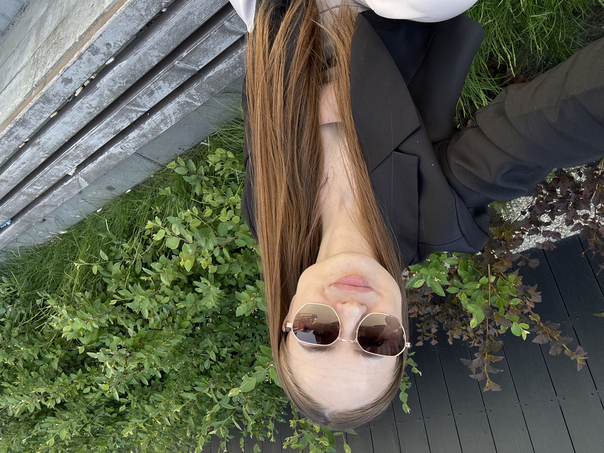 A woman with long brown hair and sunglasses is posing for a photograph. She is wearing a black blazer and white top. She is standing next to a wooden fence with green grass and shrubbery at the bottom. The image is taken in an urban environment.