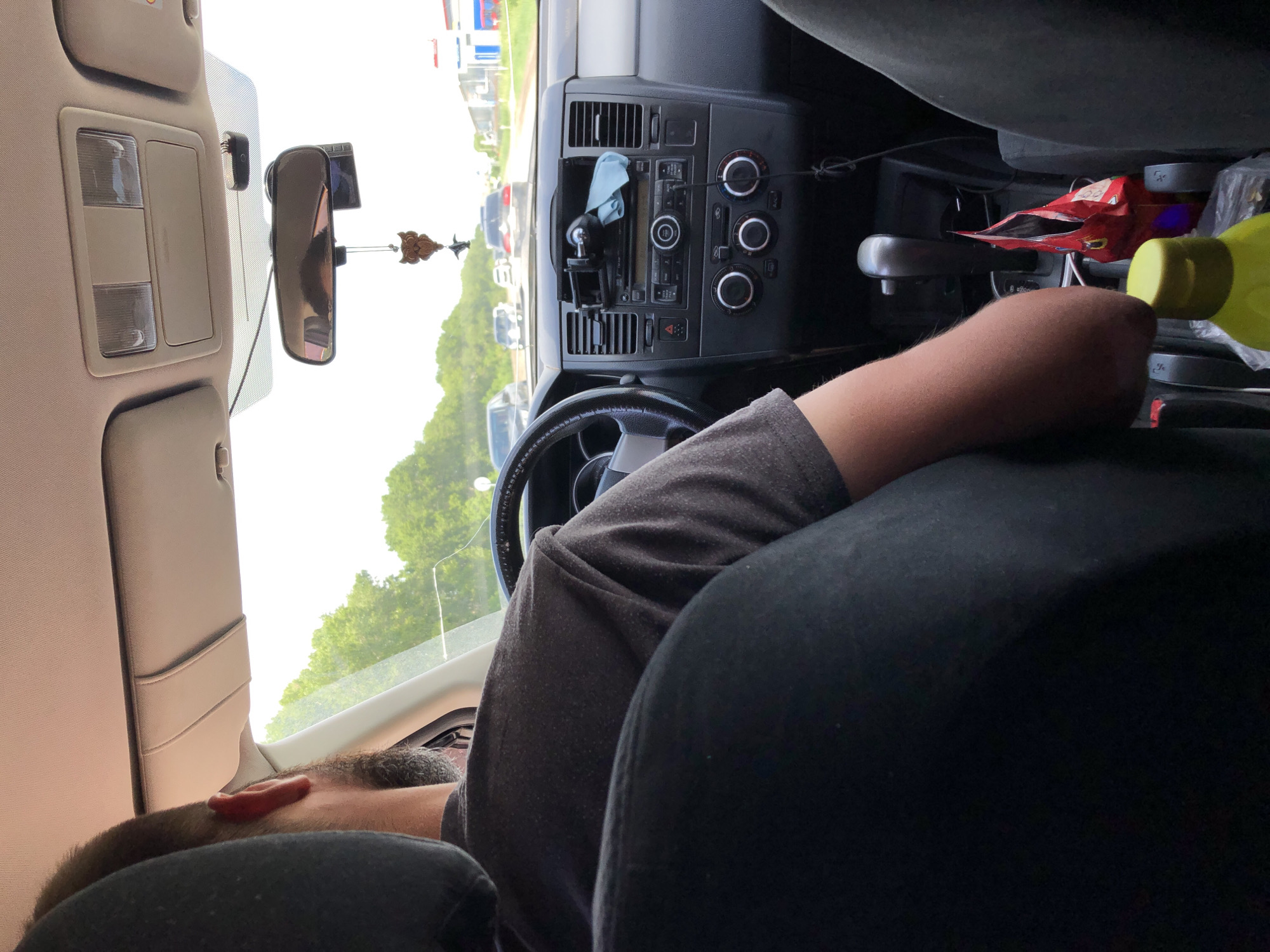 An interior shot of a car shows the back of the driver's head and torso, as well as the car's dashboard and passenger seat. The driver, a person with a beard, is wearing a dark t-shirt and has their right arm resting on the center console. The dashboard features a car stereo, climate controls, and a rearview mirror with an owl-shaped air freshener hanging from it. A phone mount is attached to the windshield. Outside the car, trees and other vehicles are visible on a road, suggesting the car is in motion in an urban or suburban environment. There is also a bag of chips and a yellow bottle in the passenger seat area.