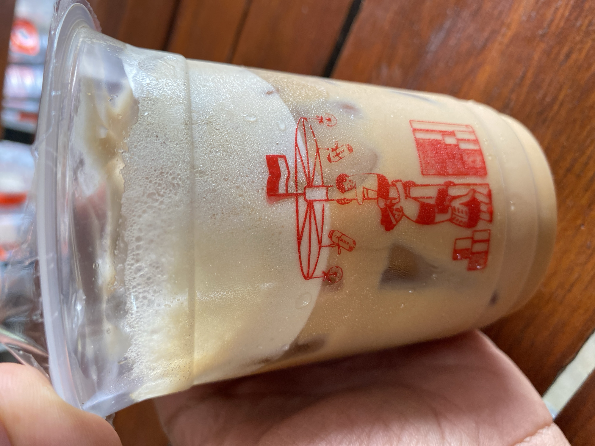 A close-up, eye-level shot shows a clear plastic cup of iced coffee with a foamy layer on top, held by a person's hand and fingers. The cup is placed on a wooden table. A red graphic design is printed on the outside of the cup, featuring a carousel-like structure with small figures and a flag. The background is blurred but suggests an outdoor setting with a wooden surface and possibly other tables. The lighting is bright and natural. The scene is likely taking place at an outdoor cafe or restaurant in Bireuenmeunasahdayah, Indonesia, given the information provided.