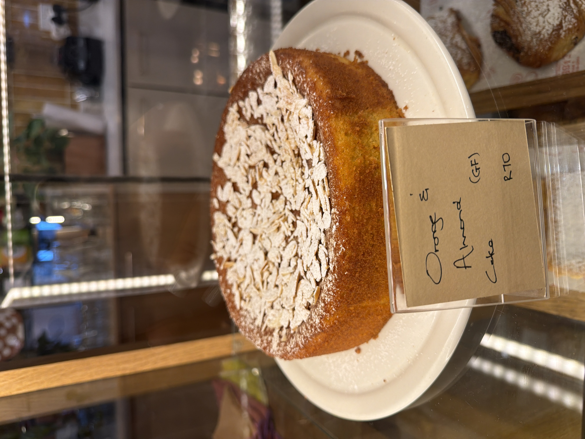 The image depicts an orange almond cake on a white plate inside a glass display case. The cake is topped with sliced almonds and powdered sugar. Next to the cake is a handwritten label that reads "Orange Almond (GF) Cake R70." The location is likely a bakery or cafe, potentially in Cape Town, South Africa, considering the price in Rands. Other pastries are partially visible in the background.