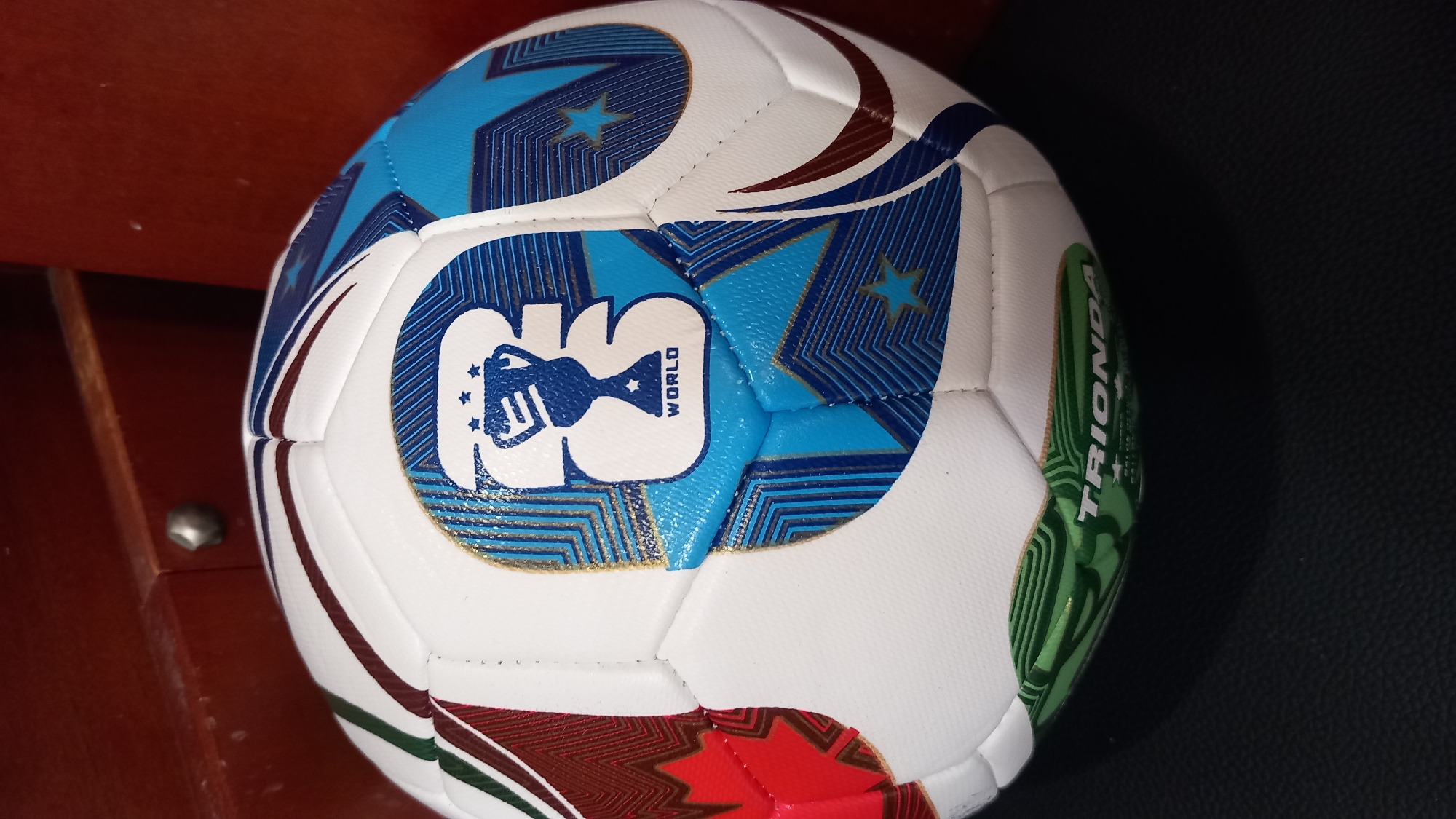 A close-up, slightly angled shot shows a white soccer ball with various colored panels and graphics. A prominent blue and white logo features a trophy with stars and the word "WORLD" in a smaller font. The ball has green, red, and brown patterned sections. Part of the word "TRIONDA" is visible on a green panel at the bottom of the ball. The ball rests on a dark, textured surface, possibly a table or floor, with a blurred reddish-brown wooden background behind it. The image does not provide any information about the city of Kos, Greece.