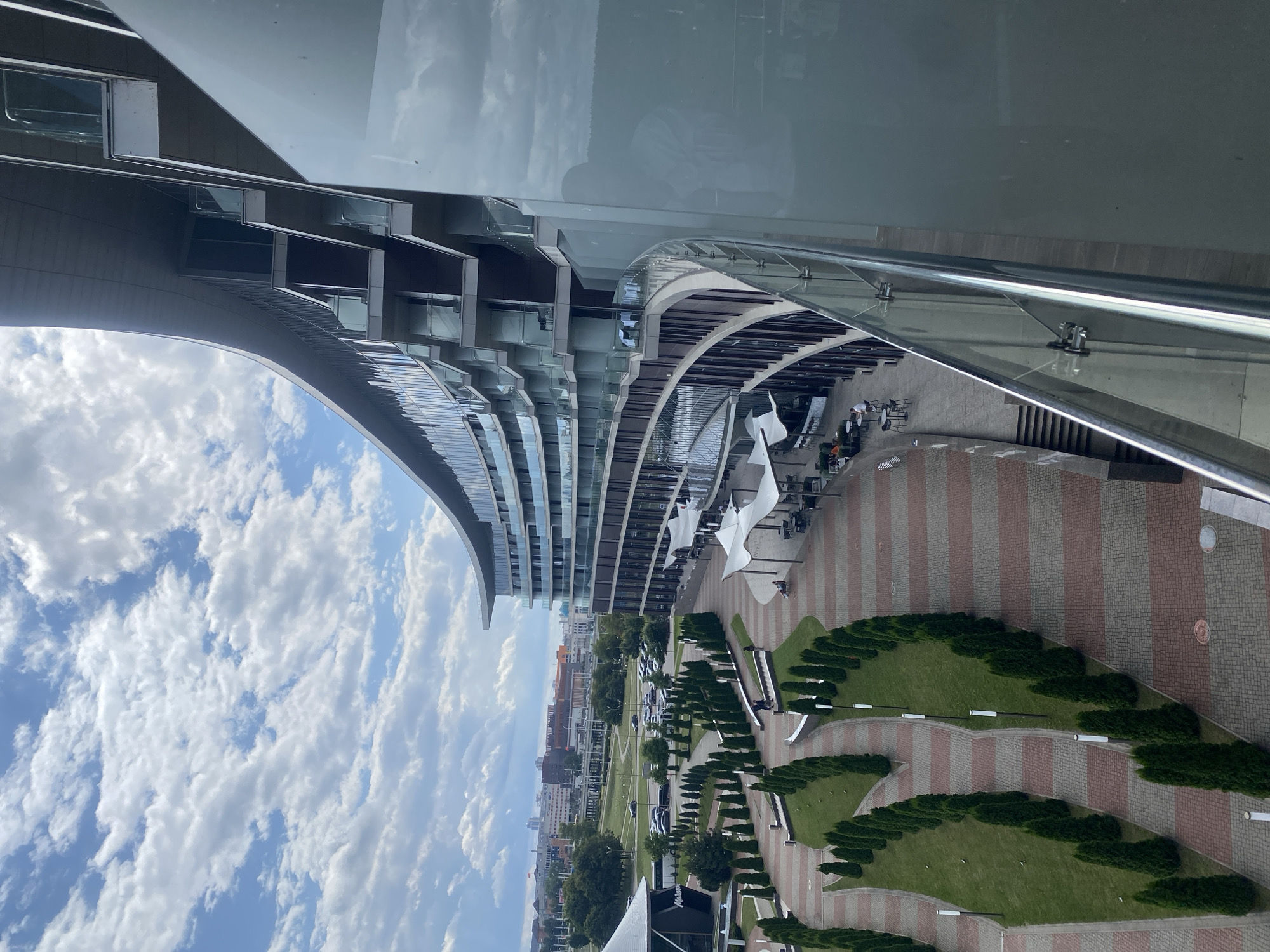The image captures an elevated view of a modern, curved building with multiple balconies, likely a hotel or residential complex. The building's facade is a mix of glass and dark vertical elements, with balconies featuring glass railings. Below the building, a landscaped plaza area is visible, with rows of manicured evergreen trees lining curved pathways. The pathways are paved with a pattern of red and grey rectangular bricks. Several white, tent-like structures are set up in an outdoor seating area, with tables and chairs beneath them. People are visible at some of the tables, and a few individuals are walking along the pathways. Beyond the plaza, a city landscape unfolds, featuring other buildings, roads, and parked cars, suggesting an urban environment. The sky above is partly cloudy with white cumulus clouds against a blue background. This scene is located in Minsk, Belarus, specifically the area near the Svislach River and the Minsk Sea, where the National Library of Belarus is situated.