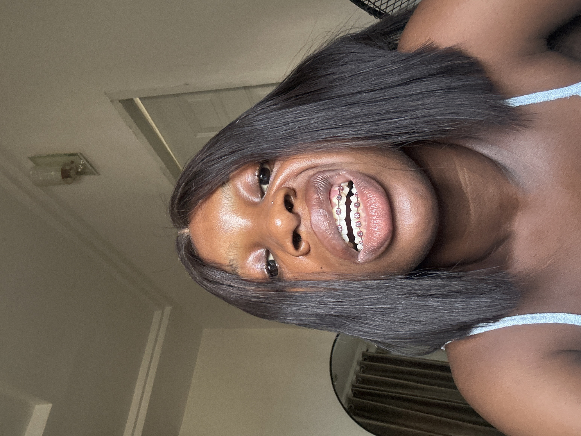 A close-up, selfie-style photograph depicts a woman indoors. Her complexion is dark brown and her hair is straight, dark brown, and shoulder-length. She is smiling, revealing teeth with pink braces. A small piercing is visible on her left nostril. She is wearing a light blue top with spaghetti straps. In the background, an off-white wall with a recessed doorway or window is visible, as is a light fixture. Behind her, a set of beige vertical blinds are visible. Based on the architectural style and the skin tone of the individual, the location is potentially in Abuja, Nigeria, but this cannot be definitively confirmed from the image alone. No interactions are visible. The environment is domestic and appears to be a room in a house or apartment.