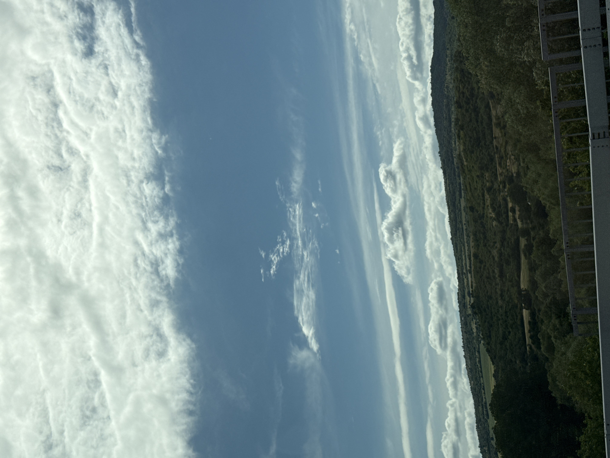 The image displays a wide-angle view of a hilly, rural landscape under a partly cloudy sky. The foreground is dominated by a grey metal railing, likely part of a bridge or overpass, suggesting the viewpoint is elevated. Beyond the railing, dense green trees and shrubs cover the lower slopes of rolling hills. Further back, the hills are less densely vegetated, revealing patches of lighter green fields and darker, more rugged terrain. The sky is a bright blue, streaked with various types of clouds, from wispy cirrus formations to larger, cumulus clouds. There are no people or animals visible in the image. The scene captures a natural, somewhat untamed environment. The provided metadata indicates the location is Blajievo, Bulgaria.