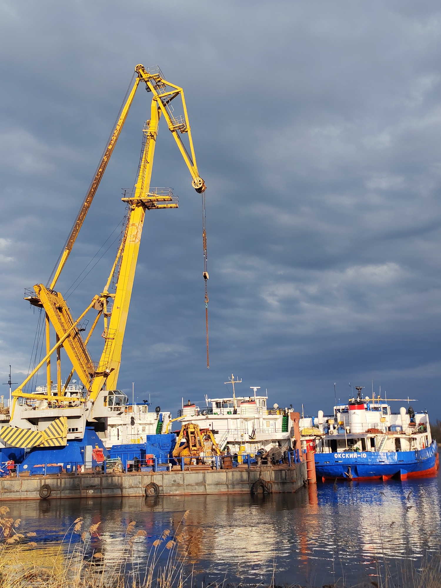 The image depicts a waterfront scene in Rybinsk, Russia, featuring a large yellow crane situated on a barge or dock. The crane is positioned prominently on the left side of the frame. Behind the crane and barge, several boats or ships are docked, including one with the name "ОКСКИЙ-10" (OKSKIY-10) visible on its blue hull. The vessels are various sizes and appear to be docked closely together. The water reflects the sky and the surrounding structures. In the foreground, there are some reeds or tall grass along the water's edge. The sky above is filled with dark, heavy clouds.
