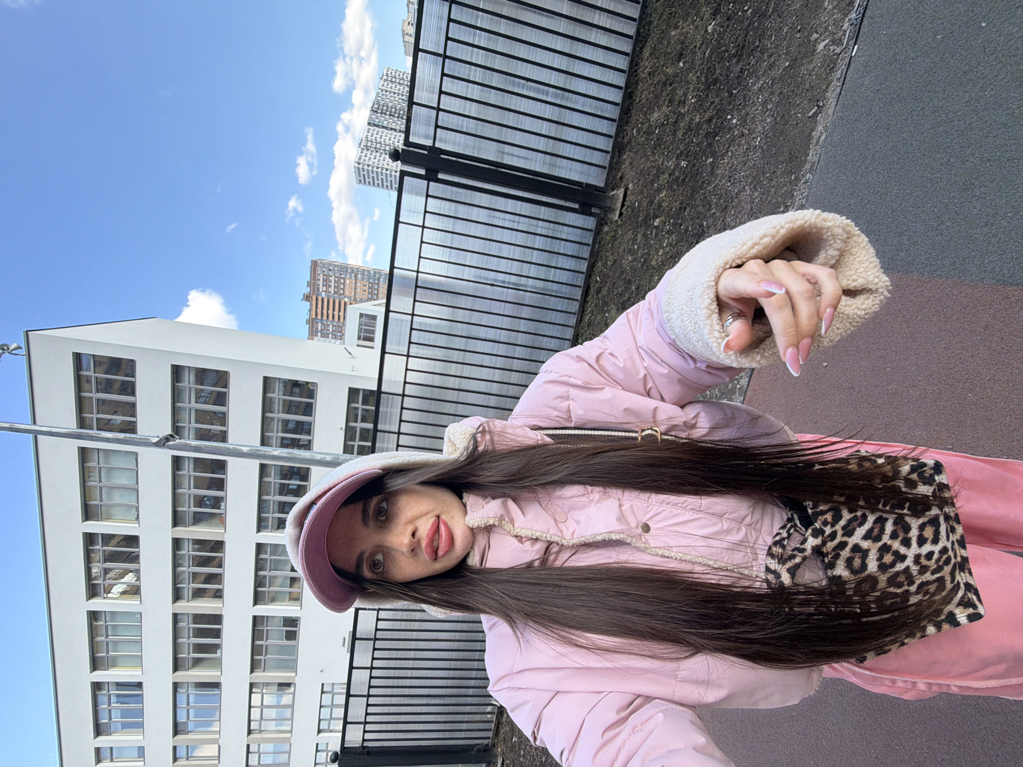 A young adult female is depicted in a selfie, positioned slightly off-center to the left, capturing herself outdoors on a clear day. She has long, straight dark brown hair, a light complexion with freckles, and wears a light pink puffer jacket featuring cream-colored sherpa lining on the collar and cuffs. Her attire includes a light pink baseball cap worn over a light-colored hooded garment and matching light pink pants. A small leopard print crossbody bag is slung over her right hip. Her left hand, adorned with a silver ring on the ring finger and French tip nails, is extended forward, presumably holding the camera. She gazes upwards and to her right.
The immediate foreground consists of a dual-toned ground surface, transitioning from dark grey to a reddish-brown, indicative of a paved area or track. Adjacent to this, a strip of dark, dry soil with sparse vegetation is visible. A black metal fence, constructed with vertical bars and translucent corrugated panels, runs horizontally across the midground. Behind the fence, a prominent, multi-story white building with numerous rows of large, rectangular windows occupies the background. This structure is identified as **School № 493, located at Komendantskiy Prospekt, 64к2, in Saint Petersburg, Russia**. In the far background, several modern high-rise residential buildings are visible against a clear blue sky with scattered white clouds, one notably featuring brown/orange facades and others with white, horizontally striped designs. A tall, thin metal pole stands vertically, partially obscuring the main white building.