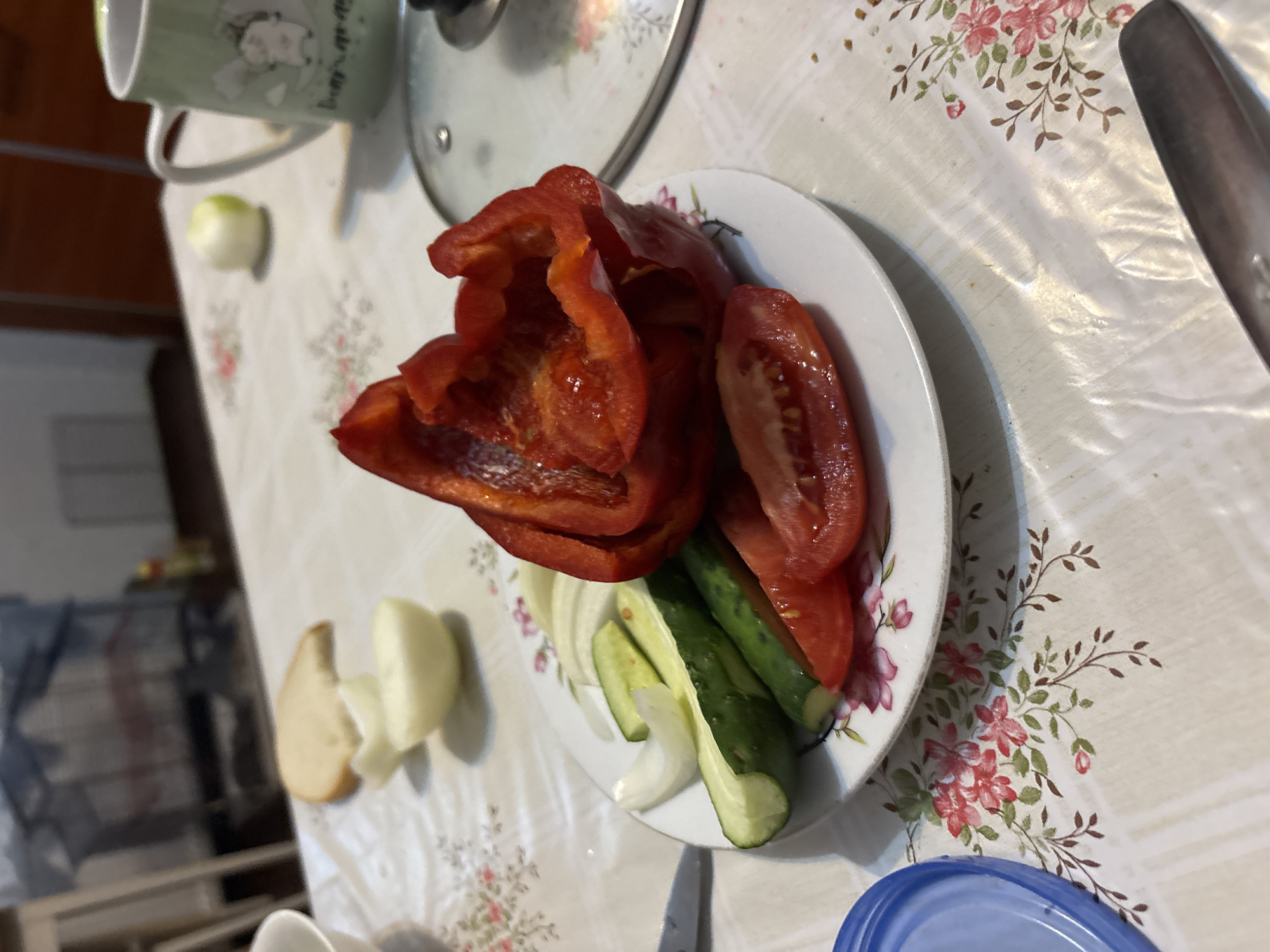 On a table covered with a floral-patterned tablecloth, a plate holds sliced red bell pepper, tomato, cucumber, and onion. A slice of bread and a whole onion are also visible on the table. A mug with a cartoon image sits to the right, and a pot lid is in the background. The scene appears to be a domestic setting, likely a kitchen or dining area, in Siemianowice Śląskie, Poland.