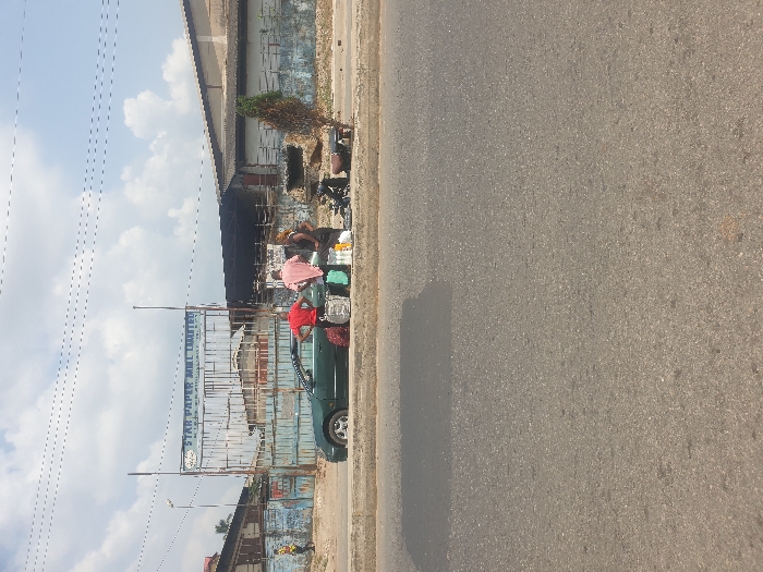 Two men are near a green car parked beside a road. One wears a pink shirt and the other a red shirt. They appear to be selling items on the street. The road surface is gray asphalt. A building with a rusted metal structure and a sign is in the background. Above the building is a cloudy sky with power lines visible. A small, dead tree stands near the edge of the road behind the men.