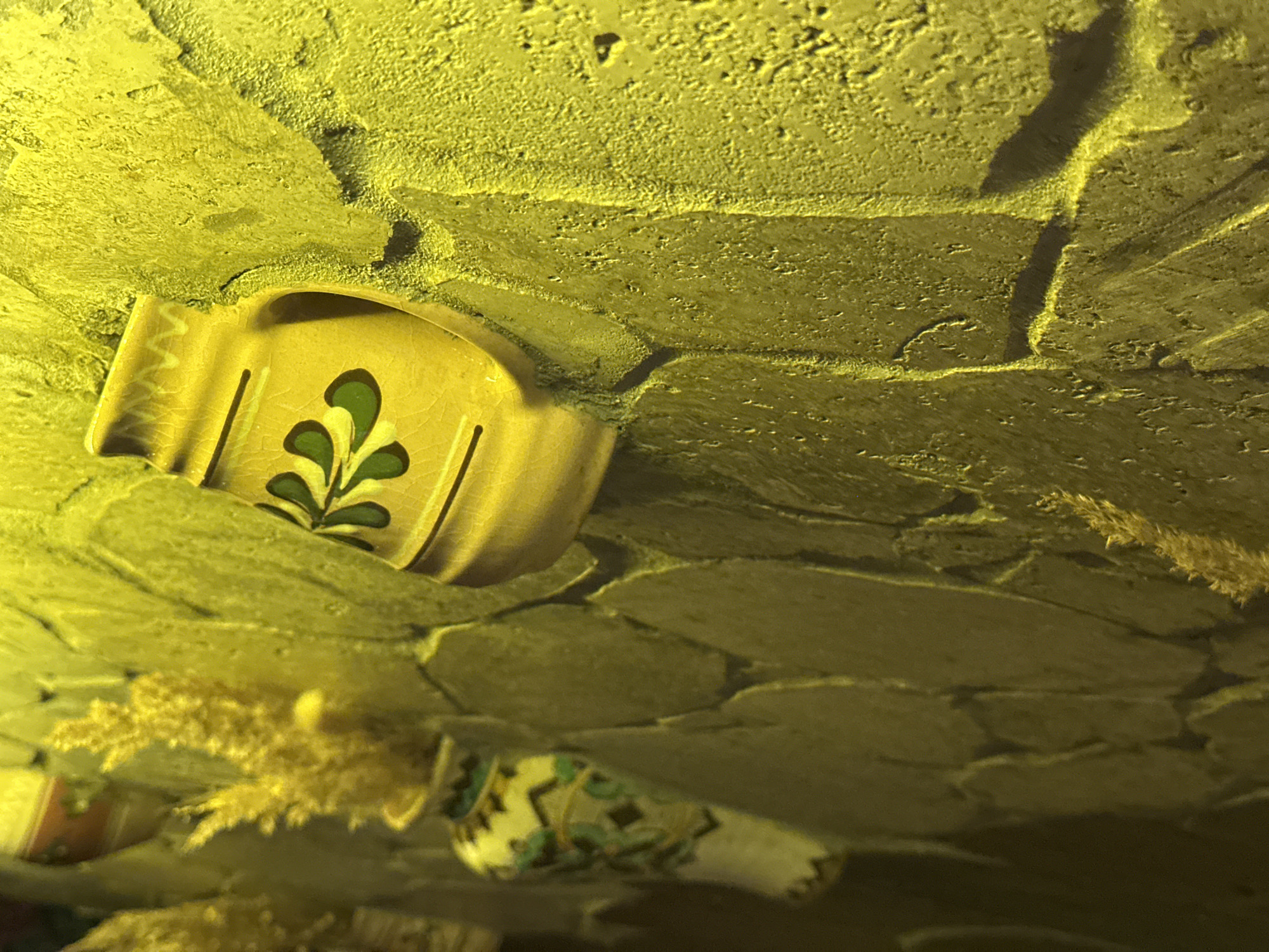 A close-up, angled shot focuses on a textured, light-yellow stone wall. Mounted on the wall are decorative ceramic items, a large yellow jug with a green floral design and a smaller, more intricately patterned vase with green and white elements. Dried floral stalks are arranged in the smaller vase. The lighting casts strong shadows, highlighting the rough texture of the wall and the glossy surfaces of the ceramics. The scene appears to be an outdoor decoration, possibly in Ternopil, Ukraine, given the context. There are no people or animals present.