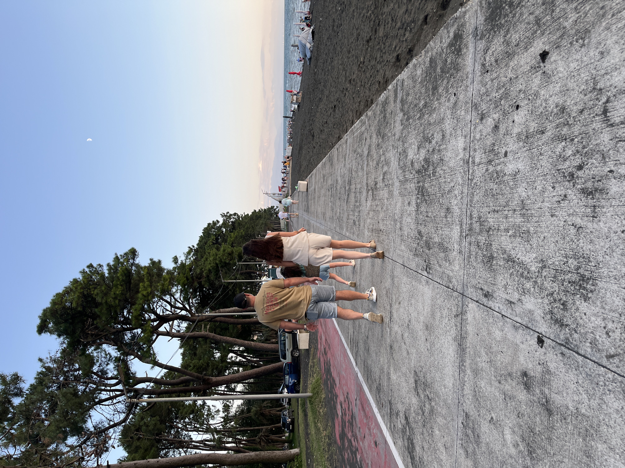 The image depicts a concrete promenade in Ureki, Georgia, bordering a black sand beach. A family of four, including two adults and two children, are walking away from the camera on the promenade. The adults are a man wearing a t-shirt and shorts, and a woman in a light-colored outfit. The children are walking ahead of them. Beyond the beach, the sea is visible with some beach umbrellas and people. In the background, there are pine trees lining the promenade, and a few cars are parked to the left. The sky is clear with a crescent moon visible.