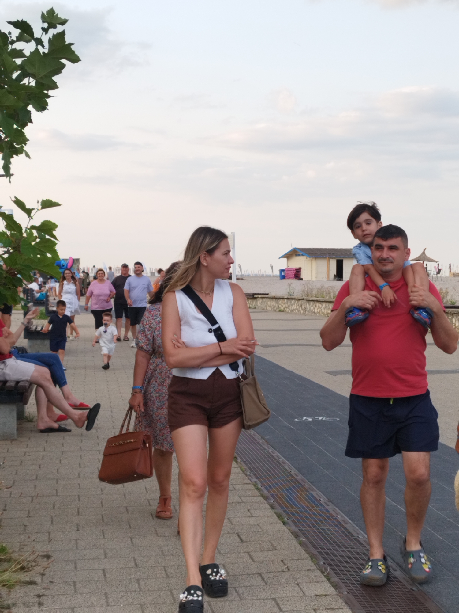 An outdoor, eye-level, full shot captures a group of people walking on a paved promenade. In the foreground, a woman with blonde hair, wearing a white sleeveless vest and brown shorts, crosses her arms and looks to the right. To her right, a man in a red t-shirt and navy shorts carries a young boy on his shoulders. The boy, wearing a blue collared shirt, looks forward. Behind them, a woman in a floral dress carries a brown handbag. Further in the background, several other people are walking, including a man in a light purple t-shirt and black shorts, a woman in a pink striped t-shirt and white shorts, a child in a dark blue t-shirt and shorts, and a toddler in a white outfit wearing a pacifier. In the far background, there is a beach with a small building and umbrellas. The scene is set in Constanța, Romania, likely along the waterfront promenade. The sky is overcast with clouds, and greenery from trees is visible on the left side of the frame.