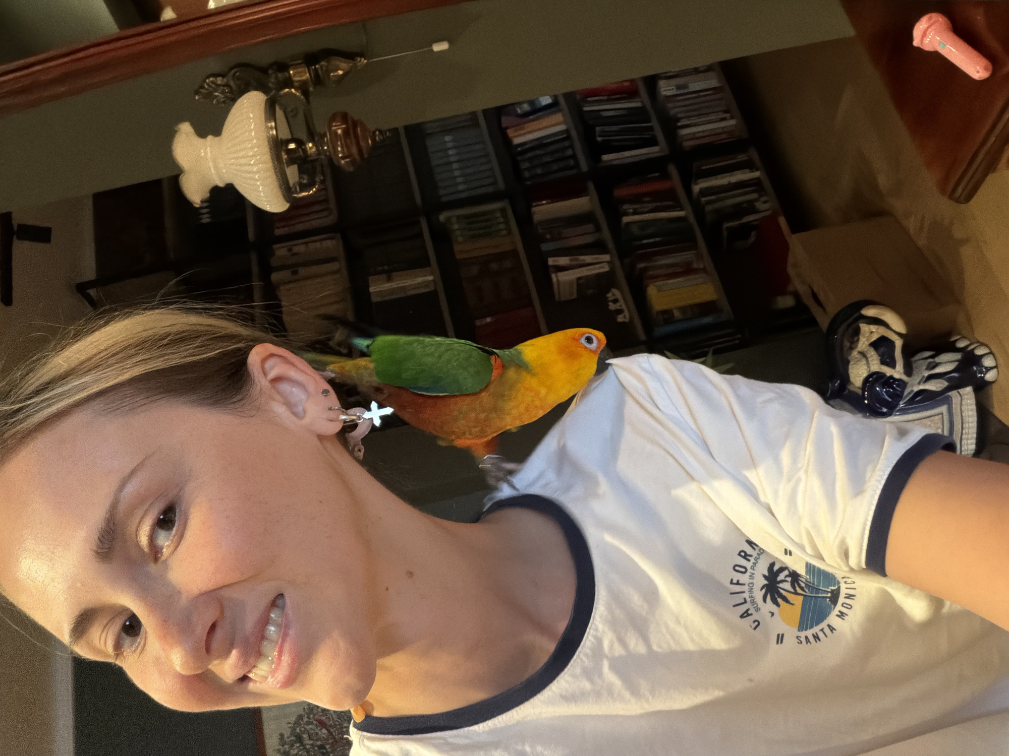 A blonde woman is captured in a close-up, selfie-style shot, her head tilted to the left and her mouth open in a smile. She is wearing a white t-shirt with a "California Surfing in Paradise Santa Monica" graphic. A small, colorful bird, predominantly yellow and green, is perched on her left ear, its head turned towards the camera. The background features a bookshelf filled with books, a vintage-style wall lamp, and a wooden table. The scene appears to be indoors. There is no information or visual evidence in the image to suggest the location is the city of Khimki, Russia.