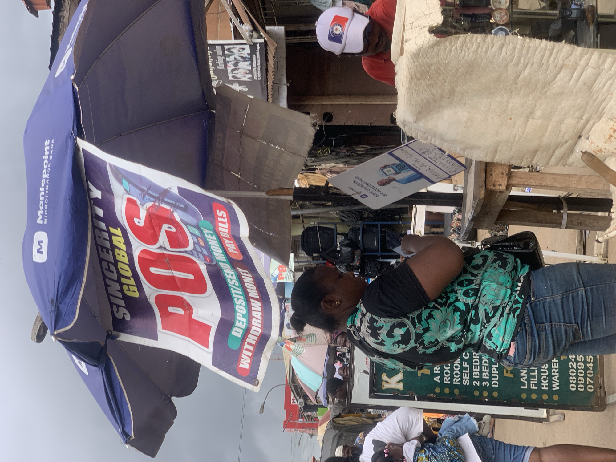The image depicts an outdoor commercial scene, likely a bustling market area in Ikorodu, Nigeria. Prominently featured is a blue and white Moniepoint umbrella advertising POS (Point of Sale) services, highlighting deposit, send money, withdrawal, and bill payment options. An African woman with an updo hairstyle, wearing a patterned green top and jeans, stands near the POS stand, her arms crossed.
To the right, a weathered wooden structure is visible, possibly a stall, with real estate advertisements in the forefront. The poster advertises apartments of different sizes and properties for sale. In the background, there is a man wearing a red shirt and a cap. Other signs and commercial displays can be seen in the distance, along with blurred figures of people walking, suggesting a crowded environment. The sky appears overcast.