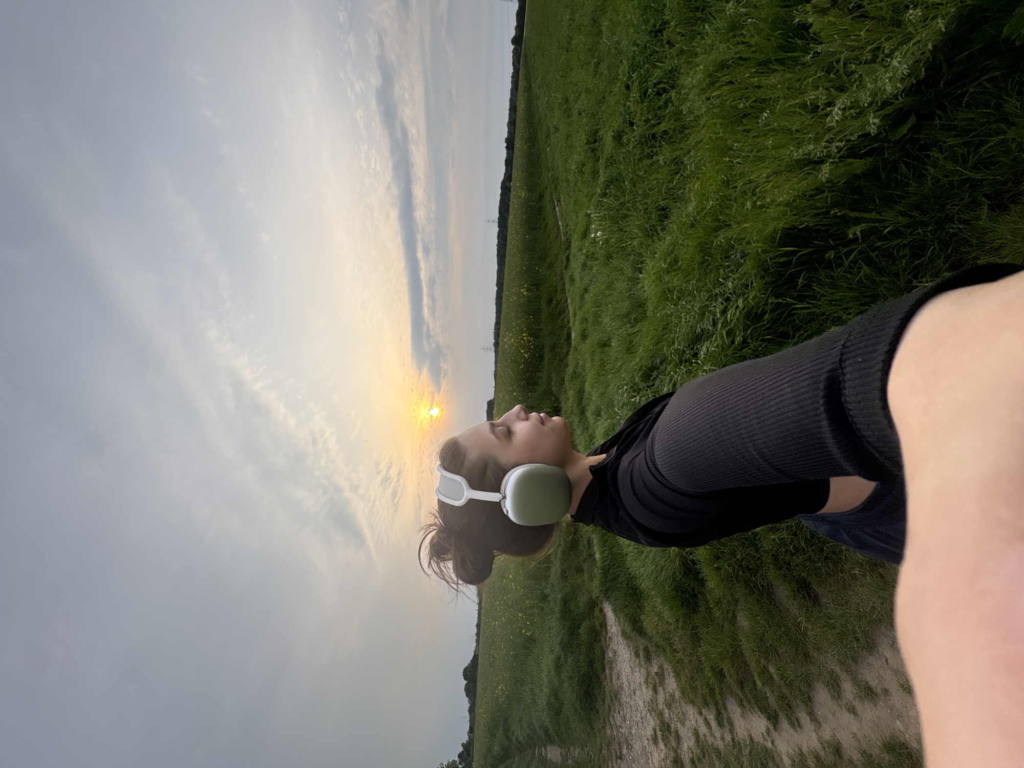 A woman is outdoors, possibly in the city of Mönchengladbach, Germany, during twilight. She is wearing white headphones and a black long-sleeved shirt. She is positioned near a field with long grass and a dirt path. The sky is overcast, with the sun visible through the clouds.