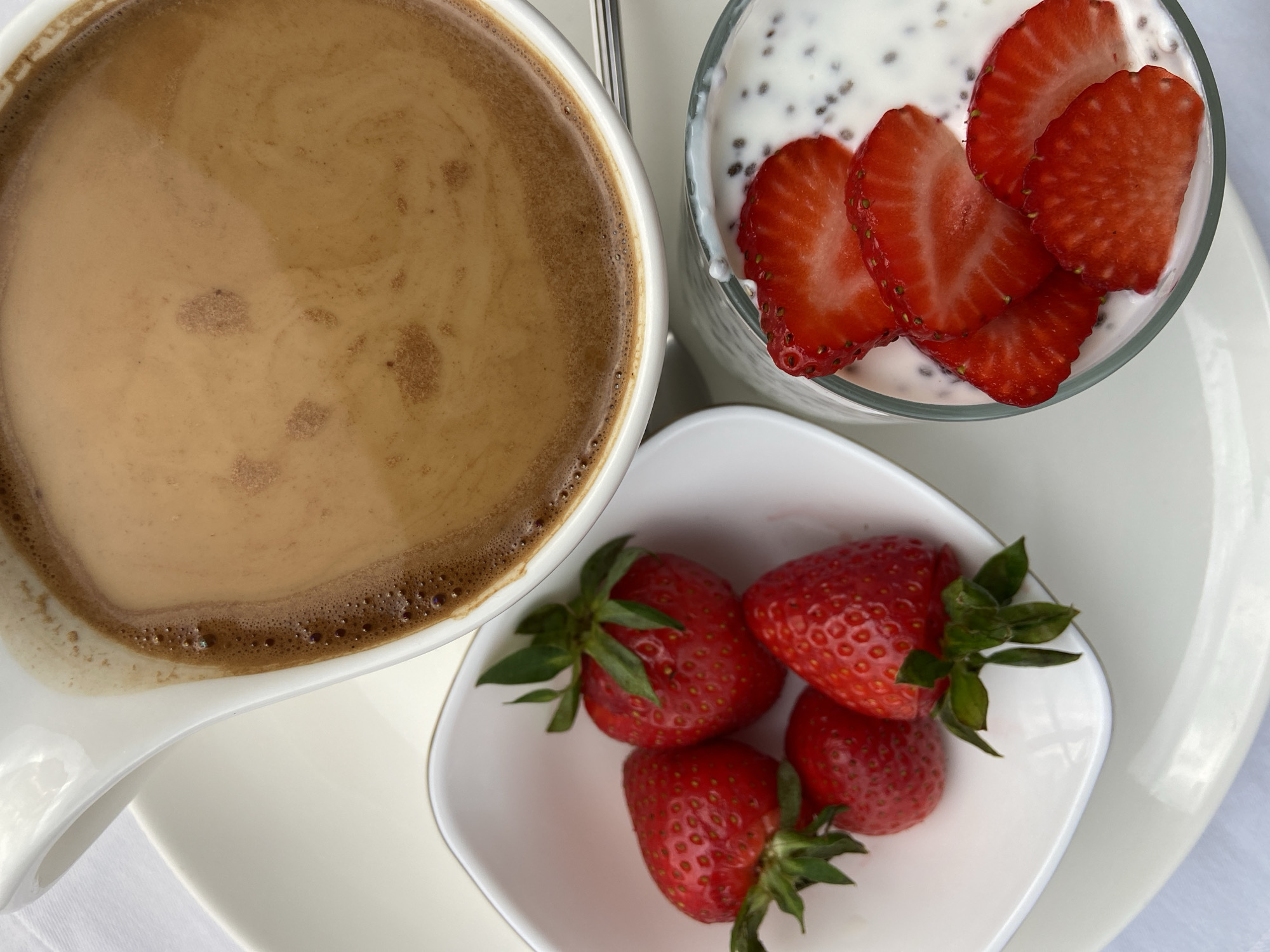An overhead shot captures a healthy and aesthetically pleasing breakfast or snack arrangement on a white surface, possibly a table, in Budva, Montenegro.
The primary subjects are a large cup of coffee, a glass of chia pudding or yogurt with strawberries, and a small bowl of whole strawberries.
On the left, a white, elegantly shaped cup holds a dark brown coffee with a layer of lighter brown crema on top, indicating it might be an espresso-based drink. The surface of the coffee shows subtle swirling patterns.
To the upper right of the coffee cup, a clear glass contains a white, creamy substance, likely yogurt or chia pudding, generously speckled with dark chia seeds. Several bright red strawberry slices are artfully arranged on top of the pudding. A metallic spoon handle is partially visible behind the glass.
Below the glass and to the right of the coffee cup, a small, square-shaped white bowl contains four fresh, whole red strawberries, each with its green stem and leaves still attached. The strawberries appear ripe and vibrant.
All items are placed on a larger white plate or saucer, which in turn rests on a white tablecloth or surface. The overall impression is one of a clean, fresh, and inviting meal. There are no people visible, nor is there any discernible activity beyond the presentation of the food. The lighting is bright and even, suggesting either natural daylight or well-lit indoor conditions. No visible text is present in the image.