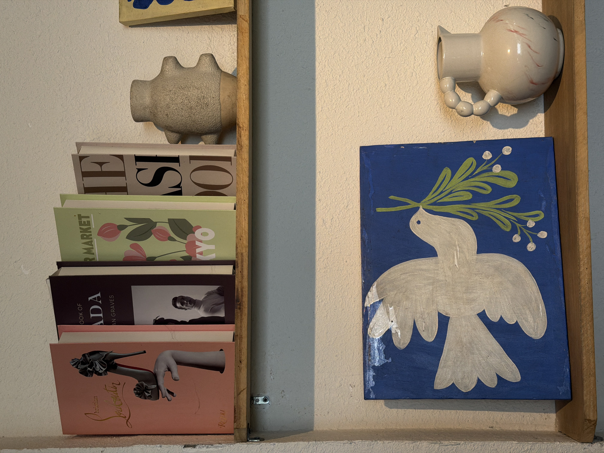 This indoor scene features a curated display of objects and books against a textured, light-colored wall. On the left, a stack of books is neatly arranged within a light wooden frame. The top partially visible book shows "THE SI". Below it, a book with a green spine reads "SUPERMARKET" and "KYO", adorned with a floral design. A dark-covered book displays "OOK OF ADA" (likely "PRADA") by "AN GRAVES" next to a black and white portrait. The bottom book is pink, featuring an illustration of a high-heeled shoe and a hand, with "Christian Louboutin" and "RIZZOLI" printed on its spine. A textured, light brown ceramic piggy bank rests on top of this book stack.
To the right, a square painting depicts a stylized white dove carrying a green olive branch with white berries against a vibrant blue background, symbolizing peace. Above the painting, a light-colored ceramic wall-mounted object, possibly a vase or holder, is visible. It has a rounded body, a beaded handle, and faint red/pink abstract markings. A wooden frame runs vertically along the left side of the book stack and partially along the right side of the painting. Small metal brackets secure the wooden frame to the wall. The scene is static, illuminated by soft, natural-looking light, creating gentle shadows. No specific time of day or weather is indicated. The image captures a decorative and calm interior setting.