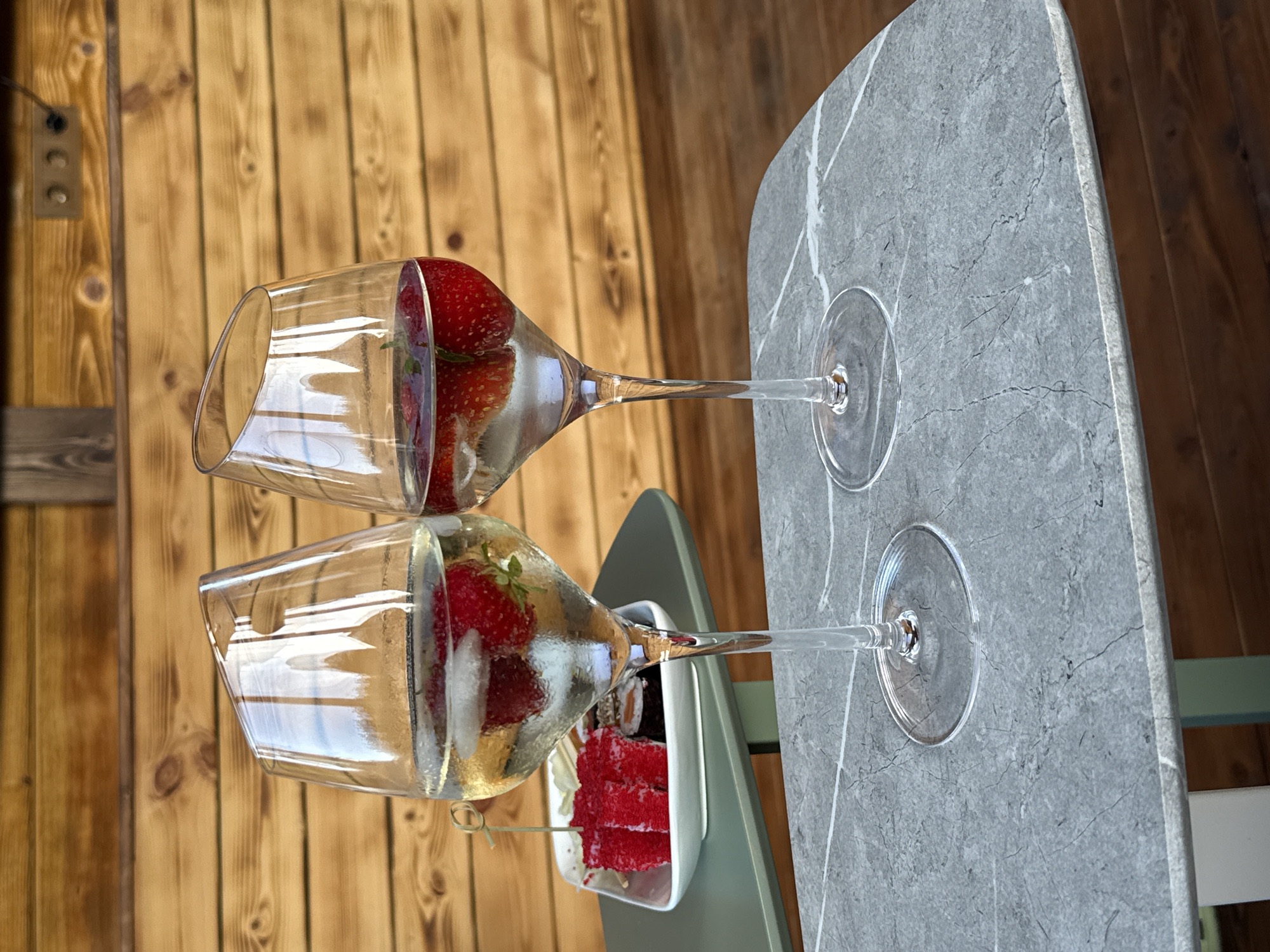 Two wine glasses filled with a clear liquid and strawberries sit on a marble-patterned table, which is placed against a wooden plank wall. In the foreground, one glass is tilted, showing the strawberries inside. Behind it, another glass stands upright, also containing strawberries and liquid. A small white dish with what appears to be sushi and red caviar is visible beneath the tilted glass. The background is a rustic wooden wall, possibly part of an outdoor setting. The lighting suggests it is daytime. No people are visible in the image.