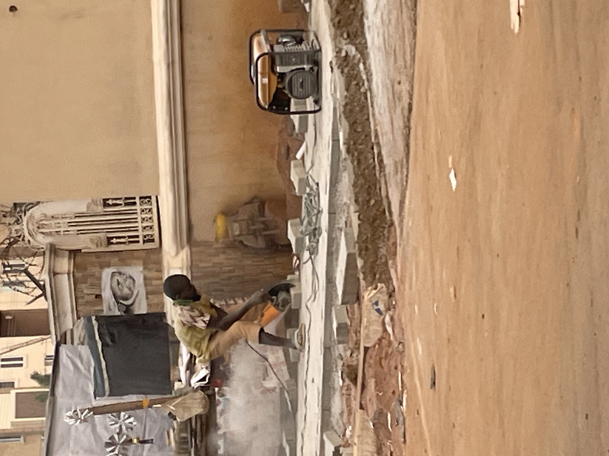 In Abuja, Nigeria, a young boy is shown cutting paving stones with a circular saw on a dirt and concrete surface. The boy, wearing a yellow and brown striped shirt and tan shorts, is positioned in the middle of the frame, focused on his work. Dust is flying from the saw as it cuts through the stone. To the right of the boy, a yellow generator sits on the ground, connected by a black cord to the saw. In the background, a building with white trim and patterned walls is visible, along with a decorative metal gate. Several stacks of grey paving stones are scattered around the work area, along with some green ropes. The overall scene depicts an outdoor construction site.