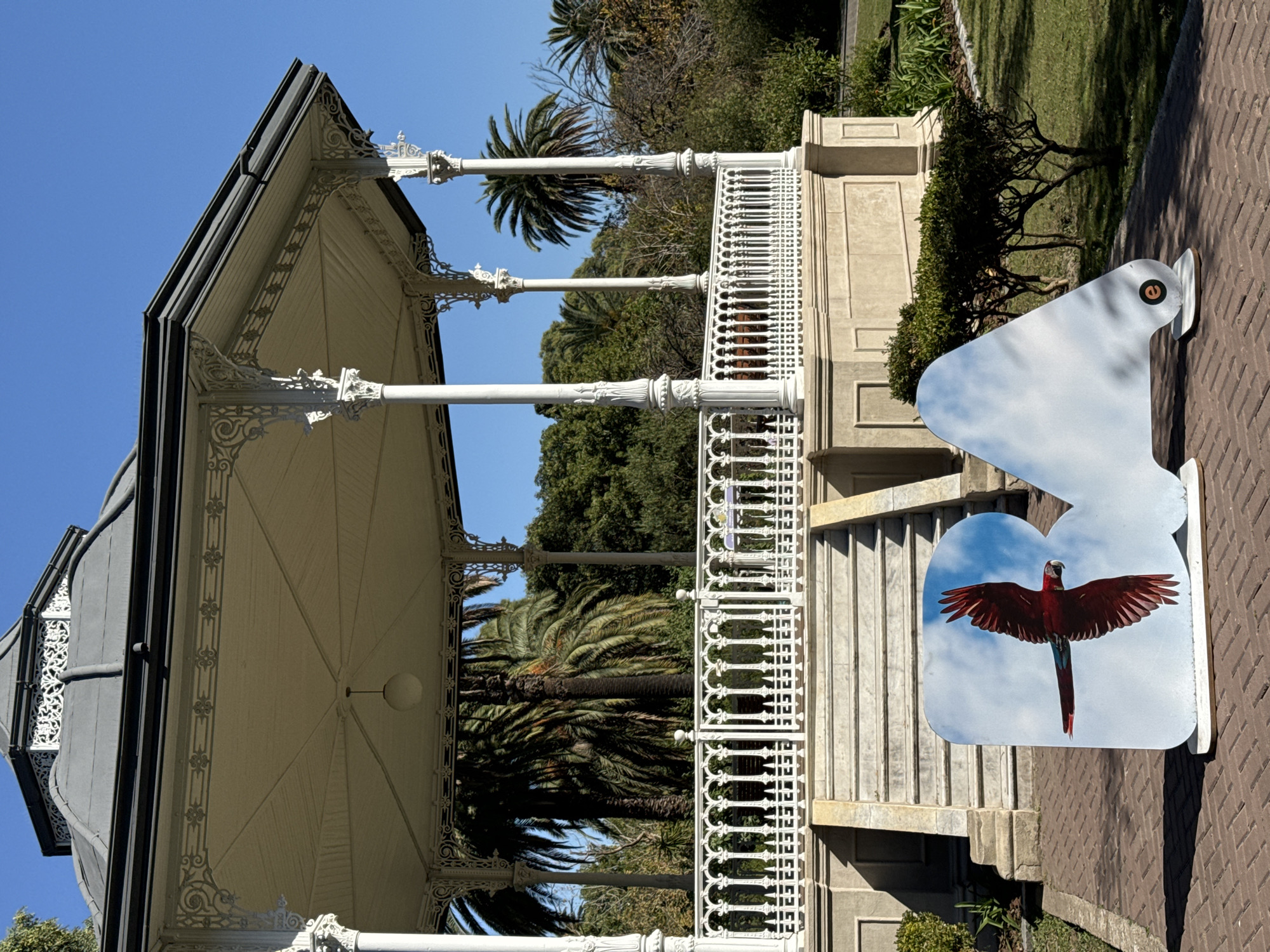 The image captures a gazebo in the Barrancas de Belgrano park in Buenos Aires, Argentina. The gazebo is white with ornate detailing and has a dark gray roof. Palm trees are visible in the background. To the right of the gazebo, there are bird-shaped informational signs on display. The ground is covered with brick pavers, and the park area consists of grass and various greenery. The sky is clear and blue.