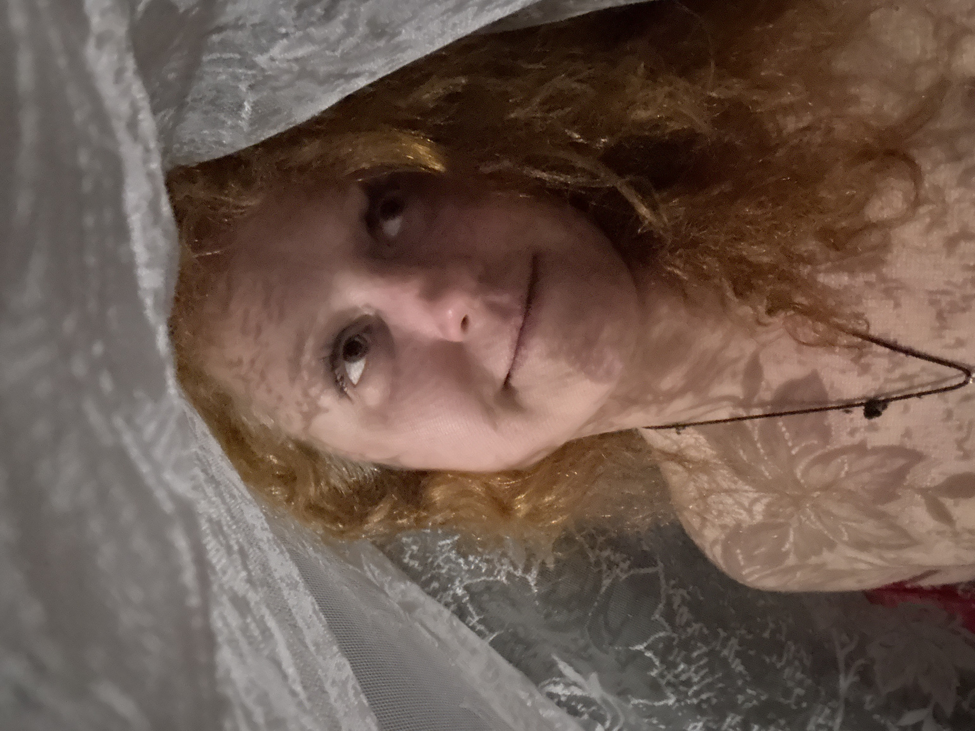 A woman with reddish-blonde hair is lying down. She is partially covered by a gray, lace-like fabric. She is wearing a thin necklace with a small, dark pendant. Her skin has a faint floral pattern on it, which is likely a shadow cast from the fabric. The photo appears to have been taken indoors. The location is not evident from the image and there is no way to determine it being in Chaikovsky, Russia.