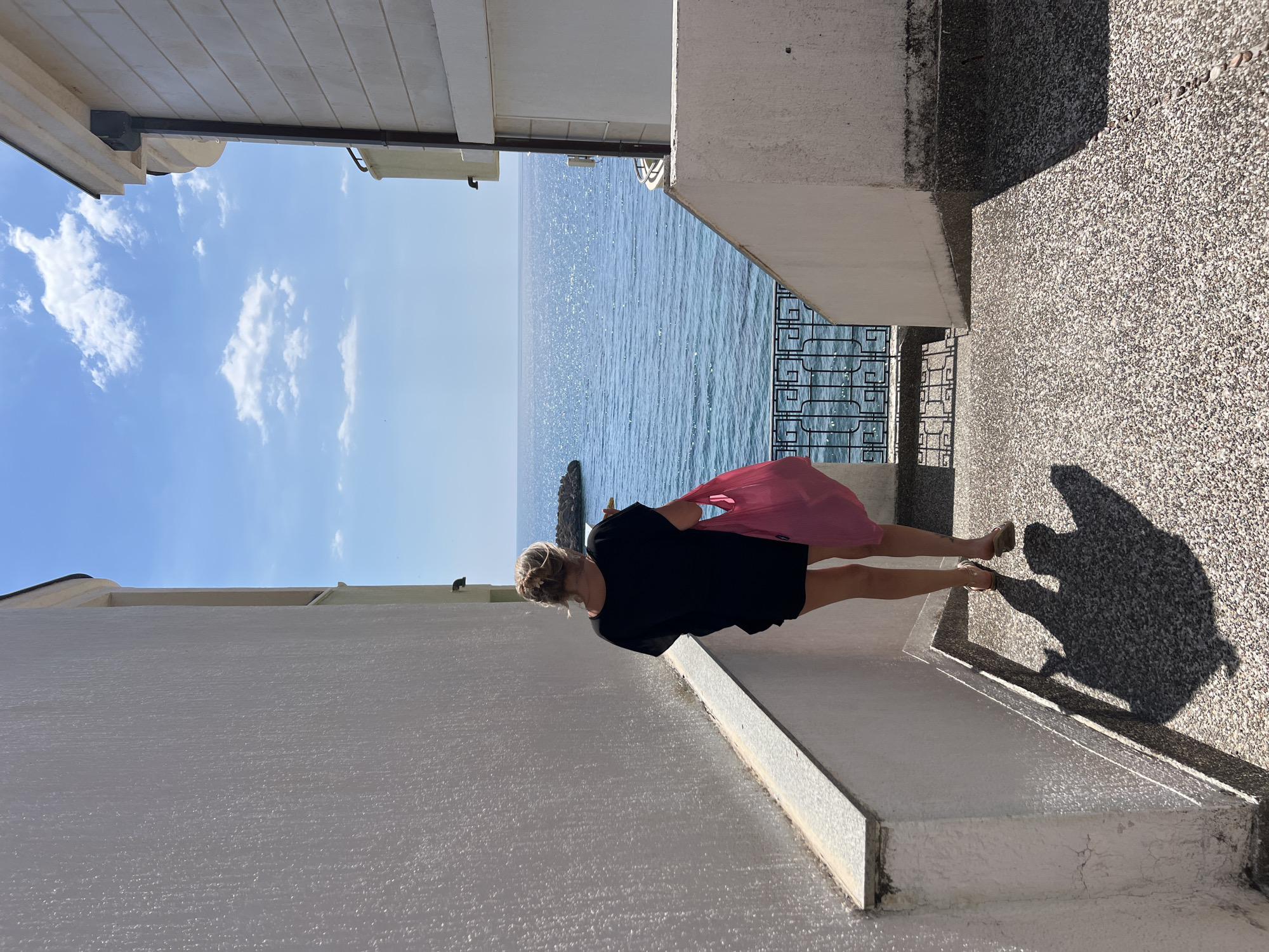 The media file depicts a woman standing on a textured pathway, holding a pink bag in her right hand. She is wearing a black dress and sandals. The woman is facing away from the camera, looking out towards a body of water. The water, likely the Adriatic Sea, is sparkling under a clear blue sky with scattered white clouds. The scene is set in Sveti Stefan, Montenegro, with glimpses of white buildings and a black metal railing visible. The sun is high in the sky, indicating it is daytime, and the weather appears to be pleasant. A large shadow of the woman is cast on the textured ground to her right. The overall mood is one of peaceful contemplation and enjoyment of the scenic view. There is no visible text in the media file.