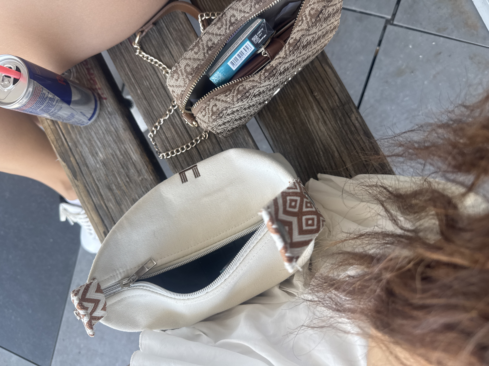 The image shows a close-up overhead view of a wooden bench with two bags, a can of Red Bull, and a person's hair. One bag is white canvas with brown and white zigzag trim and a partially open zipper. The other bag is brown and tan with a repeating pattern, a gold chain strap, and is also partially open, revealing a blue pack of cigarettes. A can of Red Bull with a pink straw stands on the bench. The background shows a paved surface and a person's legs wearing shorts and white sneakers. The scene is likely outdoors, possibly at a cafe or park. No specific location in Plovdiv, Bulgaria, can be determined from the image.