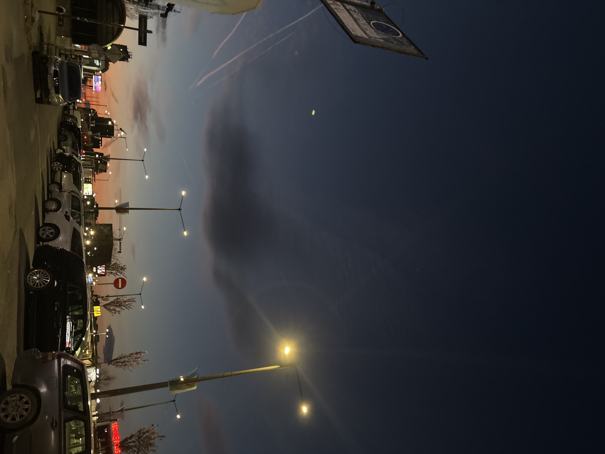 A tilted outdoor shot depicts a city scene at dusk in Sofia, Bulgaria. Streetlights illuminate a parking lot filled with cars along a commercial strip. A flag is hanging from a pole in the top right corner of the shot. The sky transitions from a muted orange-pink on the left to a deep blue on the right. Vapor trails can be seen in the sky. Trees and buildings are visible in the background. The foreground is dominated by parked cars and lampposts.