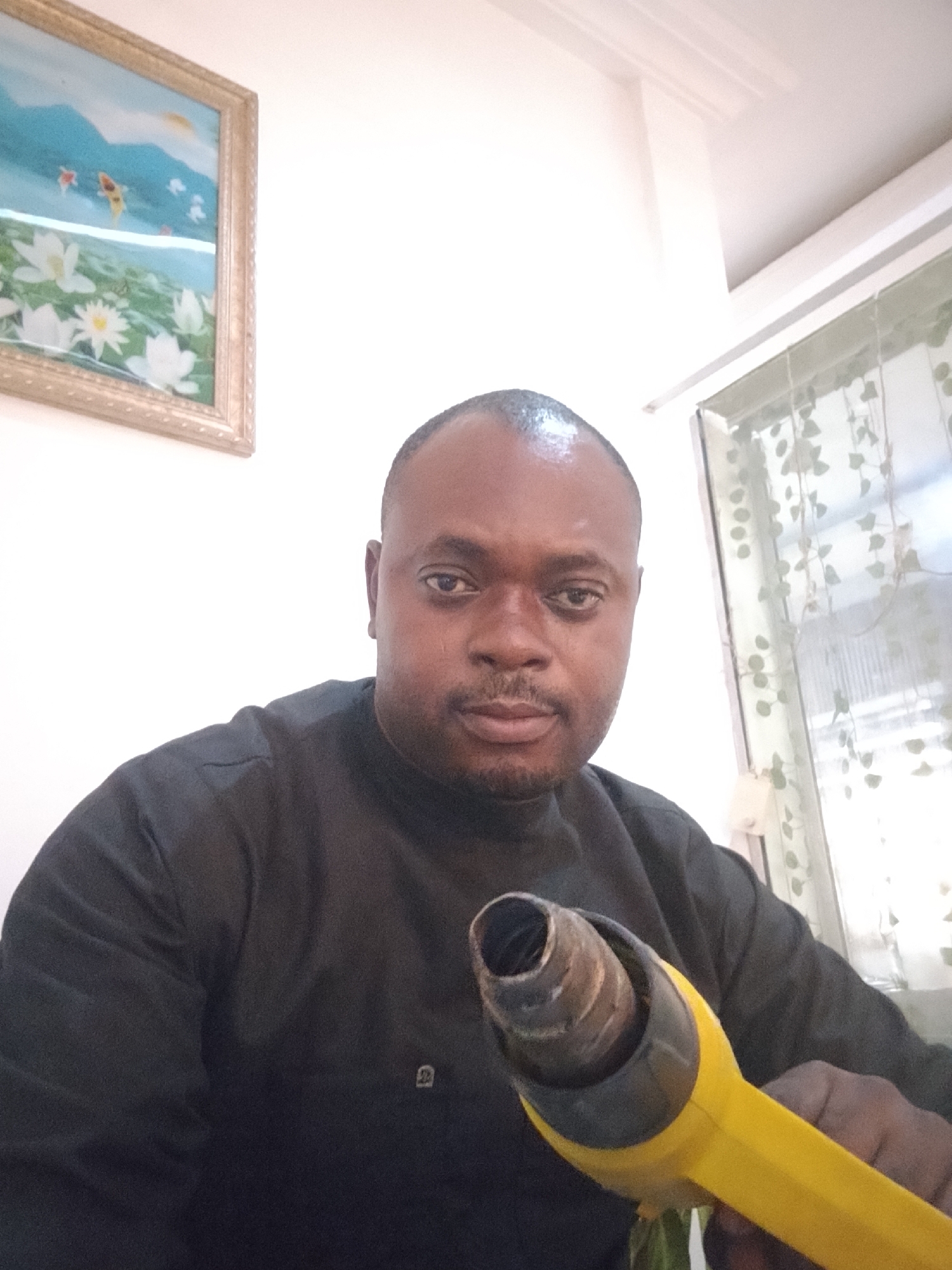 A medium indoor shot shows a man holding a yellow heat gun, likely in a residential location in Gwarinpa, Nigeria. The man has a shaved head, dark complexion, and is wearing a dark-colored shirt. He faces the camera with a neutral expression. He holds the heat gun in his right hand, its nozzle pointed toward the camera. The room has white walls and what looks like a window covered with sheer curtains in the upper right. A framed picture hangs on the wall on the left side, showing a mountain landscape.