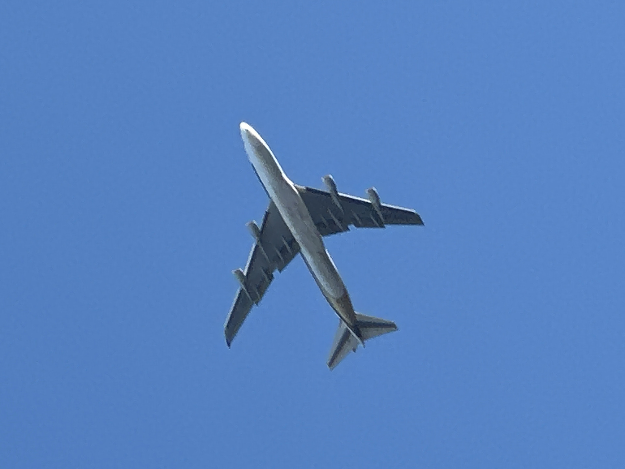 An airplane is captured from below, flying against a clear blue sky. The aircraft is a four-engine jet with its wings and fuselage prominently visible. The plane is angled, with its nose pointing upwards and slightly to the right of the frame. The underside of the wings reveals multiple engines. The tail section, including the horizontal and vertical stabilizers, is also clearly visible. The overall lighting suggests it is daytime. No specific landmarks or city details are discernible in the image, therefore, a location in London, United Kingdom cannot be determined.