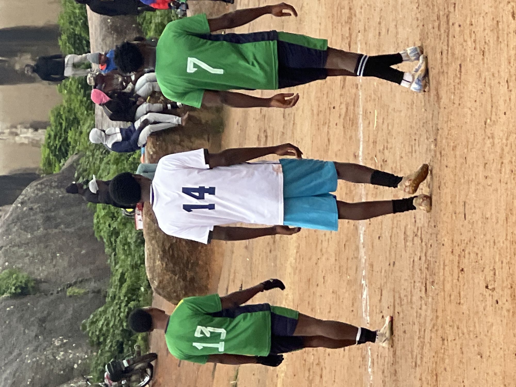 In the city of Jos, Nigeria, three young men in athletic attire walk across a dusty, uneven field. The man in the center wears a white t-shirt with the number "14" in navy blue on the back and bright blue shorts. To his left, a man in a green shirt with the number "13" in white and navy blue shorts walks away from the camera, wearing black gloves. To the right, another man in a green shirt with the number "7" in white and navy blue shorts walks in the same direction, his socks striped in black and white. In the background, a large, rocky outcrop is visible, covered in green vegetation. Other individuals are seated and standing near the rocks, and a motorcycle is parked to the left of the frame. The overall scene suggests a casual sporting event or gathering in an outdoor, natural setting within Jos.