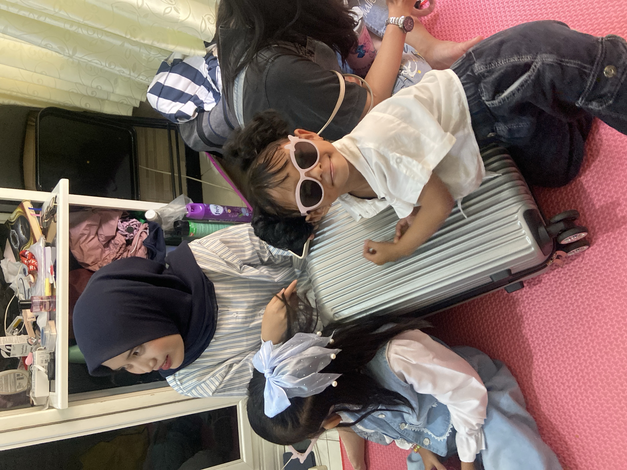 The image shows a group of people in a residential setting, specifically in Jember, Indonesia.
A woman wearing a dark blue hijab and a striped shirt is looking down towards the left. A young girl with a blue hair ribbon is situated near her. Another girl wearing pink sunglasses and a white shirt is resting on a silver suitcase. Behind her, a partially visible person is wearing a striped shirt and a dark t-shirt.
The setting is indoors, with a white shelf visible on the left, filled with various items. The floor is covered with a pink mat.