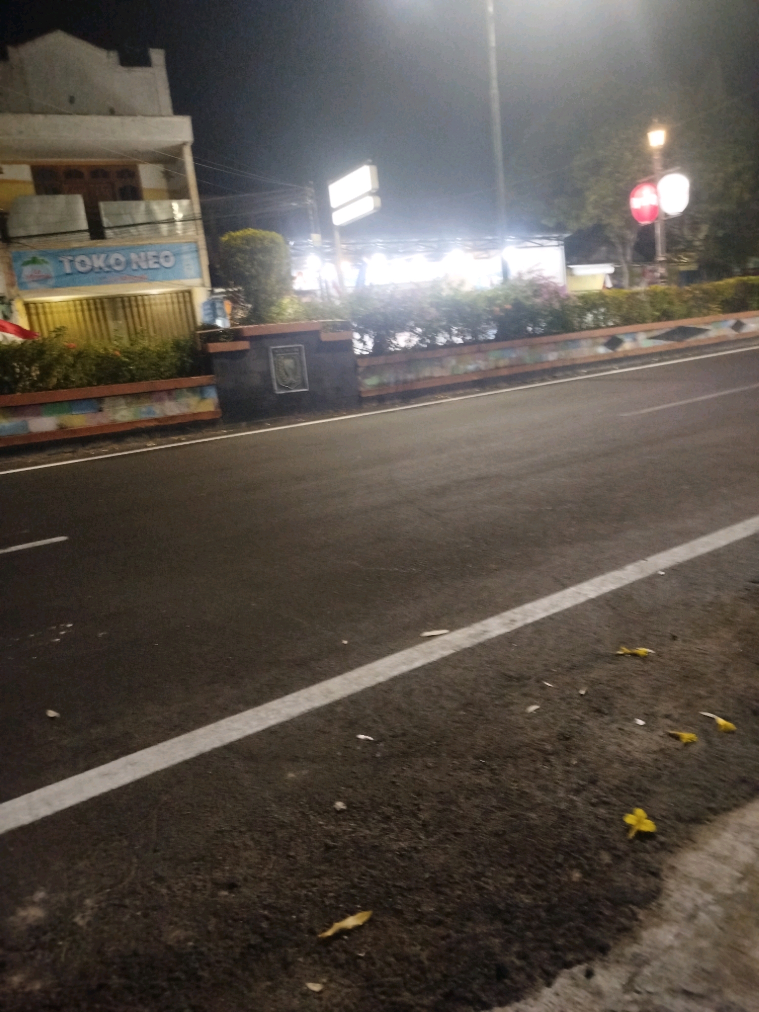 The image shows a street scene at night in Madiun, Indonesia. A building with a sign that reads "TOKO NEO" is visible on the left. The street is paved and has a white line marking its edge. A low wall decorated with colorful patterns runs along the side of the street, separating it from a landscaped area with bushes and some trees. A traffic light with red and white lights is visible in the distance. The overall impression is of a quiet street, possibly in a commercial or residential area, at nighttime.