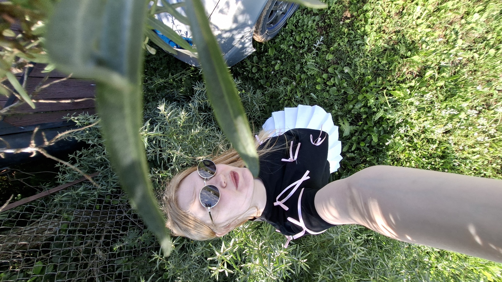 An eye-level, high-angle outdoor shot shows a young Caucasian female with long blonde hair, wearing round, mirrored sunglasses and a black shirt with pink ribbon accents and a white pleated skirt. She is lying on the ground, surrounded by green grass and plants. A portion of a vehicle is visible to the right of the frame, and a chain-link fence and wooden structure are in the background. The lighting suggests it is daytime. The location is identified as Bolshoy Bukor, Russia.