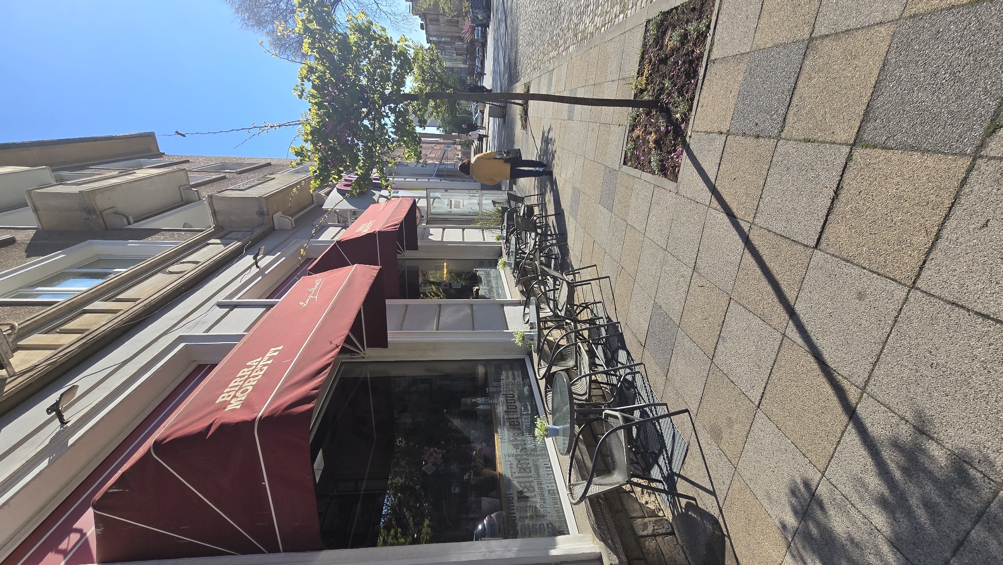 The image depicts a street scene in Stara Zagora, Bulgaria. A woman wearing a yellow jacket is walking on the sidewalk. A building with a red awning advertising "Birra Moretti" houses a business with outdoor seating. The sidewalk is paved with square tiles and lined with small garden plots. The sky is blue and the sun is shining. The scene appears to be during daytime.