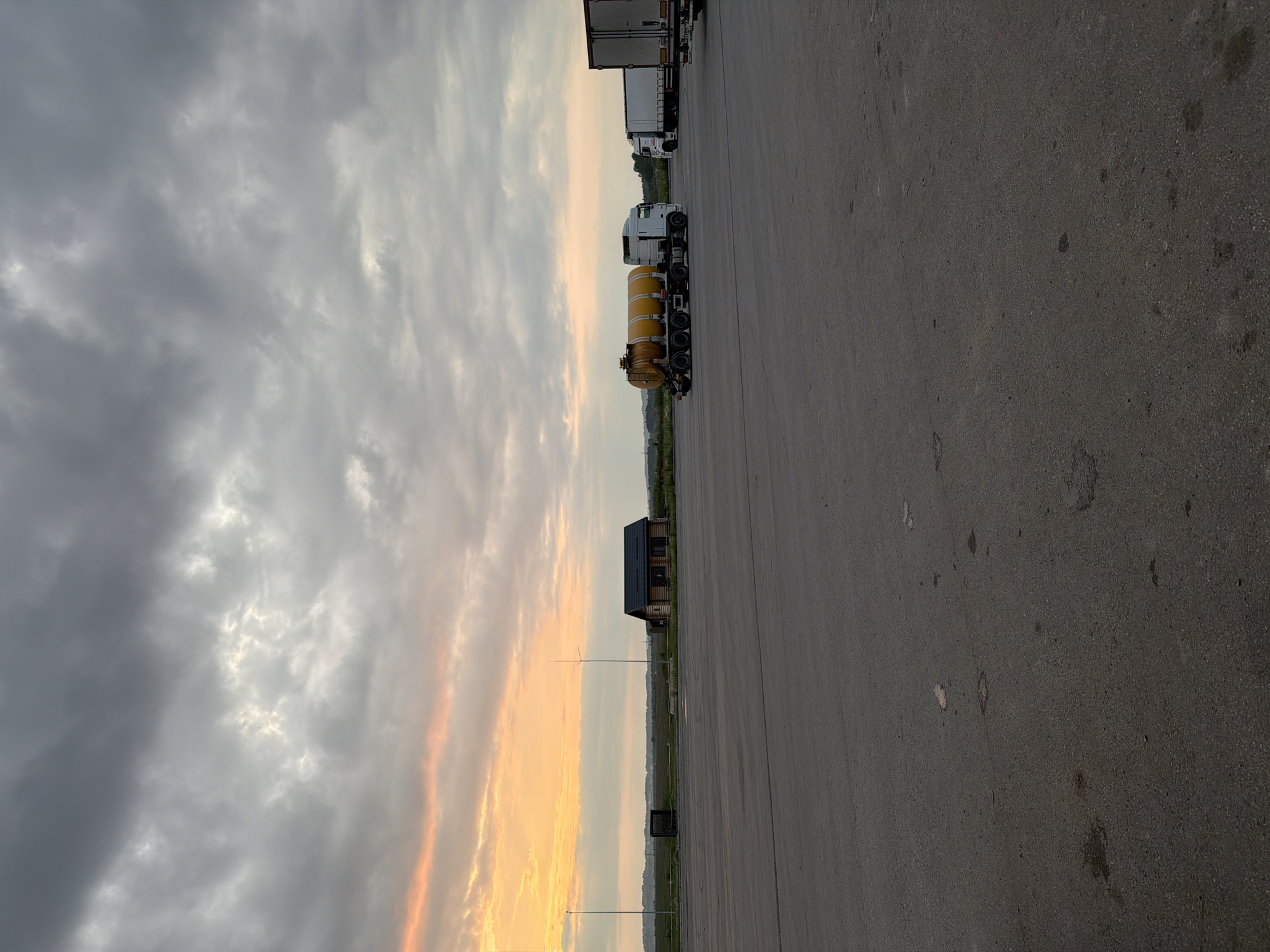 The image depicts an outdoor scene at dusk, likely in a semi-rural or industrial area, possibly near Усть-Сыны, Russia, given the provided context. The foreground is dominated by a large, empty asphalt lot. In the middle ground, to the left, a small, wooden building with a dark roof is visible, flanked by tall poles. Further to the right, several semi-trailer trucks are parked. One truck in the center has a distinctive yellow cylindrical tank. The sky above is filled with dramatic, heavy clouds tinged with the orange and yellow hues of the setting sun, particularly on the left side of the frame. The overall atmosphere is overcast and quiet, with no people or visible activity other than the parked vehicles.