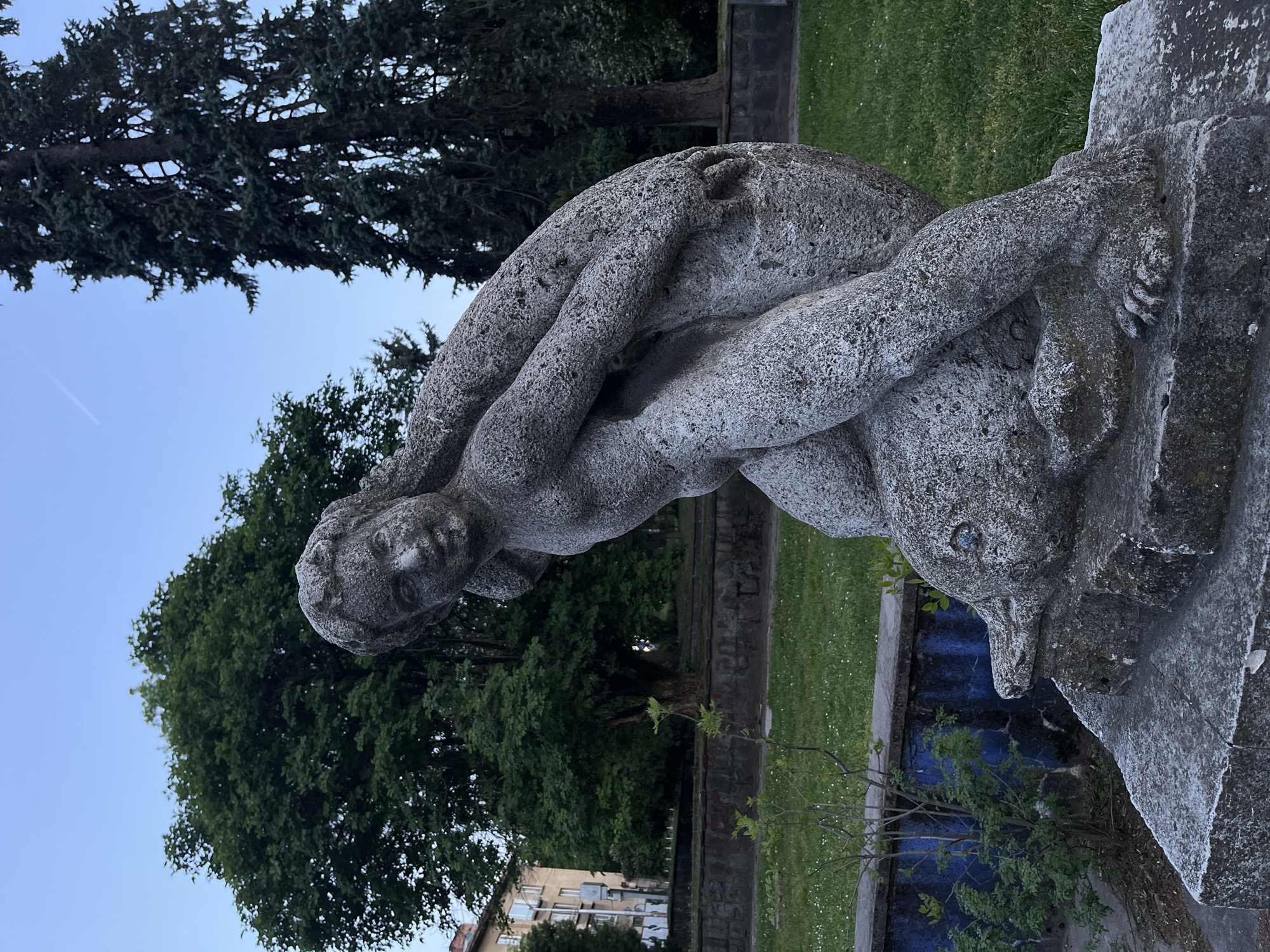 The image displays a weathered stone sculpture situated in an outdoor park, likely in Sofia, Bulgaria. The central subject of the sculpture is a human-like figure, appearing nude, captured in a bending posture. Its arms are wrapped around the head and neck of a marine animal, which resembles a dolphin or a large fish. The human figure's head is turned slightly, facing forward, while the animal's snout points downwards. Both figures are crafted from textured, aged stone or concrete, showing signs of weathering.
The sculpture is set against a backdrop of a verdant green lawn and an array of trees, including dark evergreen conifers and lighter deciduous varieties. A low wall or structure is visible behind the sculpture's base, featuring a prominent section with a vibrant blue finish, possibly painted or tiled. In the distance, a building with light-colored walls can be seen through the trees. The sky is a clear, pale blue, indicating either a late afternoon or early evening light. The scene is bathed in soft, natural illumination, conveying a peaceful and serene atmosphere within an urban park or garden. There is no visible text in the image.