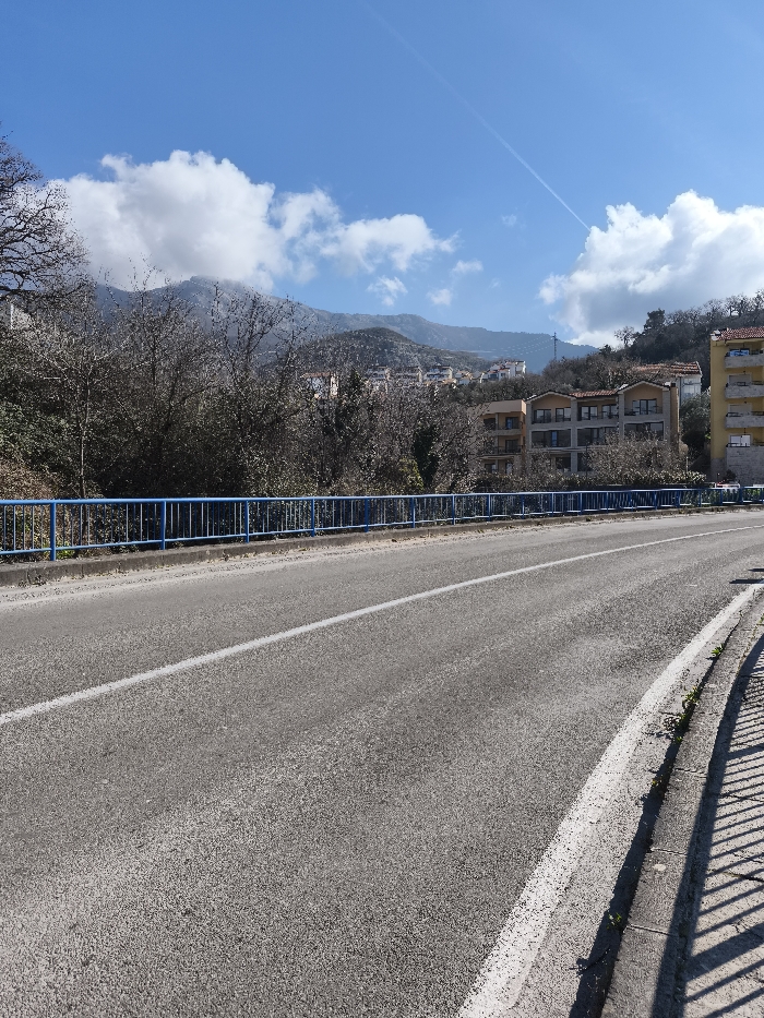 The image depicts an outdoor scene in Divanovići, Montenegro, under a clear blue sky with scattered white cumulus clouds. A faint white contrail is visible in the upper right quadrant of the sky. The foreground is occupied by an asphalt road with a visible solid white lane line. To the right of the road, a concrete curb separates the road from a sidewalk, where distinct shadows of an unseen vertical structure are cast. A blue metal barrier with multiple horizontal rails runs parallel to the road on the left. Beyond the barrier, dense, leafless deciduous trees and brush occupy the midground, suggesting a dormant season. Behind this vegetation, multi-story buildings are visible, including a yellow building with a red-tiled roof on the far right and a light-colored building with multiple dark windows to its left. In the background, large mountainous terrain rises, with some peaks partially obscured by clouds. No individuals or active subjects are present in the scene.