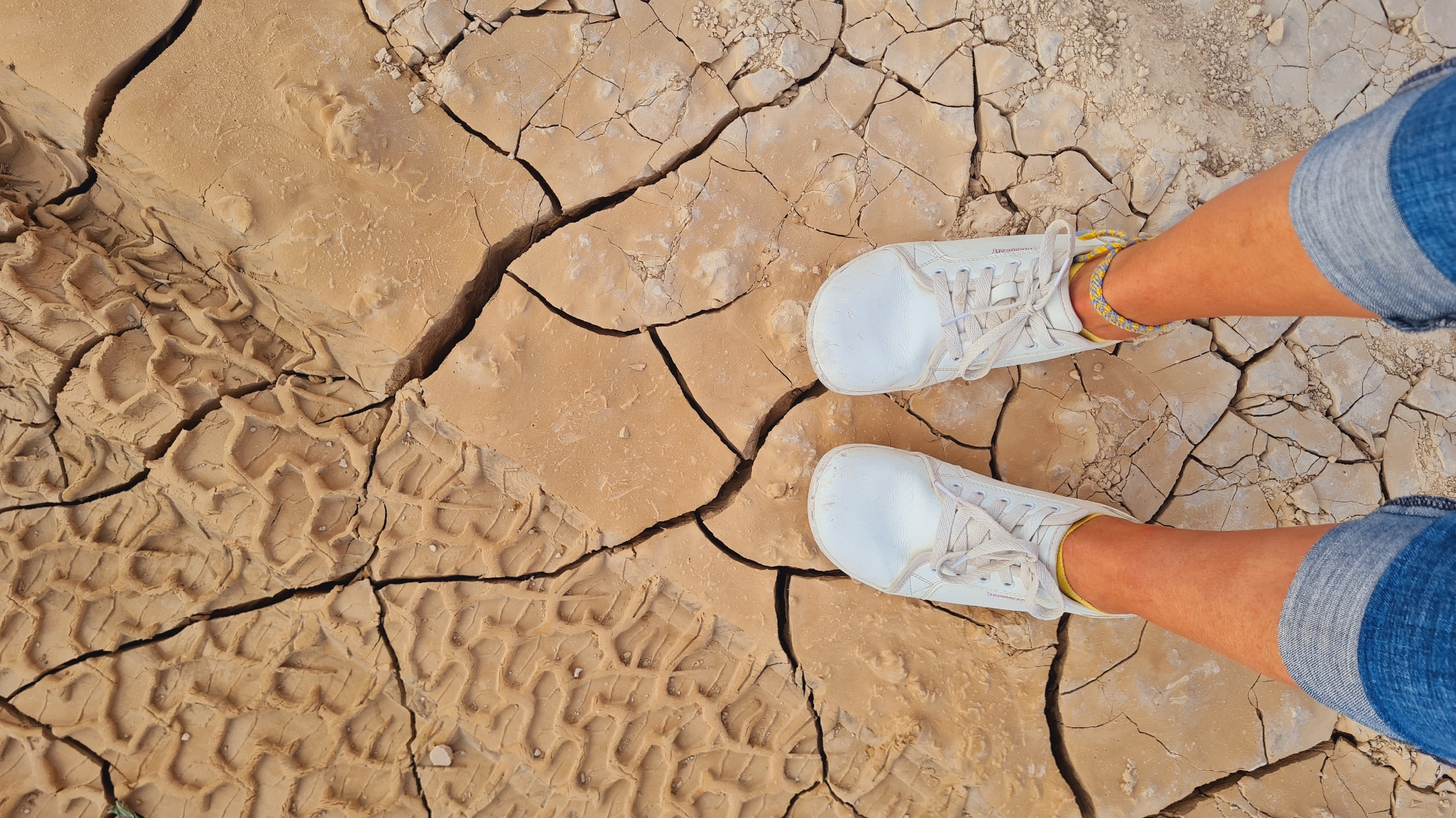 The image shows a top-down view of a person standing on cracked earth, presumably in the Tamar Regional Council, Israel. The ground is arid, with large polygonal cracks defining discrete patches of dry soil. A pattern of tire tracks is visible in the lower left of the frame. The person in the frame is wearing white shoes and cuffed denim shorts or jeans. An ankle bracelet is visible on one ankle.