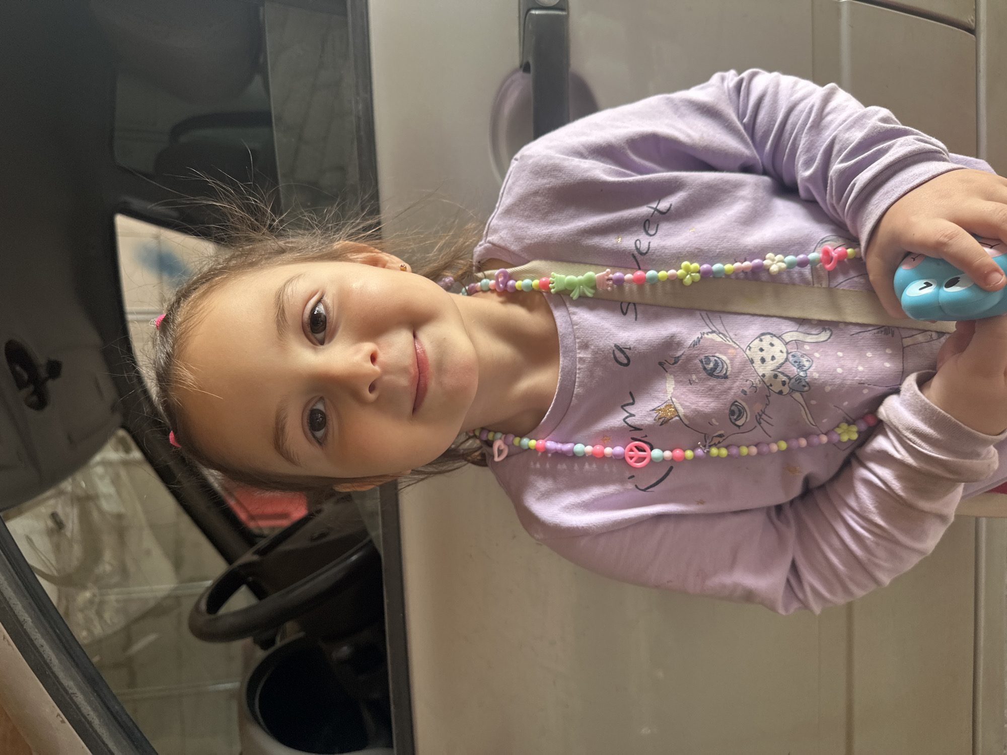 A young girl with brown hair tied in pink ponytails stands in front of a car. She is wearing a light purple long-sleeved shirt with a graphic of a cat wearing a crown and the words "I am a sweet" printed on it. She also has a colorful beaded necklace with various charms, including peace signs, flowers, and frogs. The girl is holding a light blue toy camera in her hands. The background appears to be a garage or similar indoor space, with the car's steering wheel and dashboard visible. The lighting suggests it is daytime. The girl is looking directly at the camera with a slight smile. There are no other people visible in the image.
