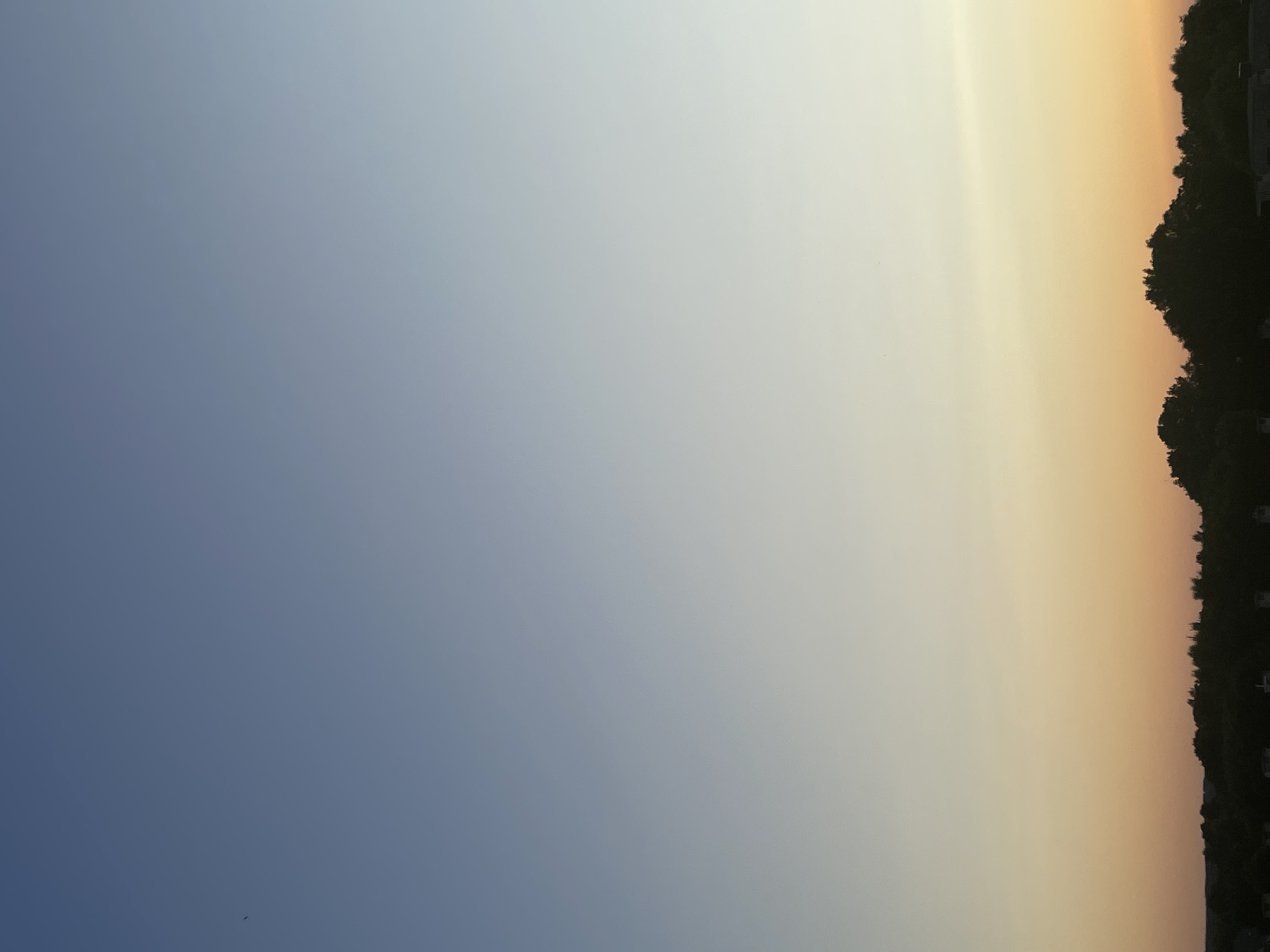 The image captures a serene sunset over Tartu, Estonia. The sky transitions from a deep blue at the top to a soft gradient of lighter blues, whites, and finally, warm hues of orange and yellow closer to the horizon. A silhouetted line of trees marks the foreground, suggesting a natural or park-like area within or on the outskirts of the city. There are no people or specific objects clearly discernible in the scene, with the focus being on the atmospheric beauty of the sky and the dark, tree-lined horizon. The overall impression is one of peaceful tranquility at the end of the day.
