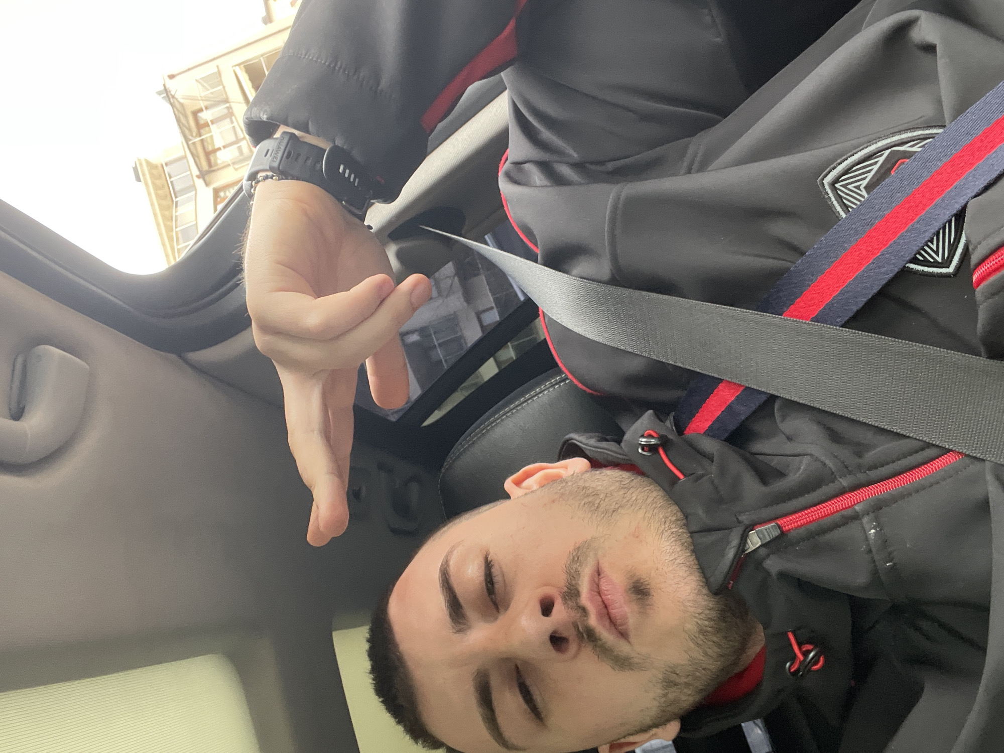 A man with a dark beard and short dark hair is seated in a car, wearing a dark jacket with red trim and a seatbelt fastened across his chest. His eyes are closed. His left hand, adorned with a dark-colored smartwatch, is raised towards the car's interior ceiling. The background visible through the car window suggests an urban environment. Due to the context clues provided, the location is Sofia, Bulgaria.