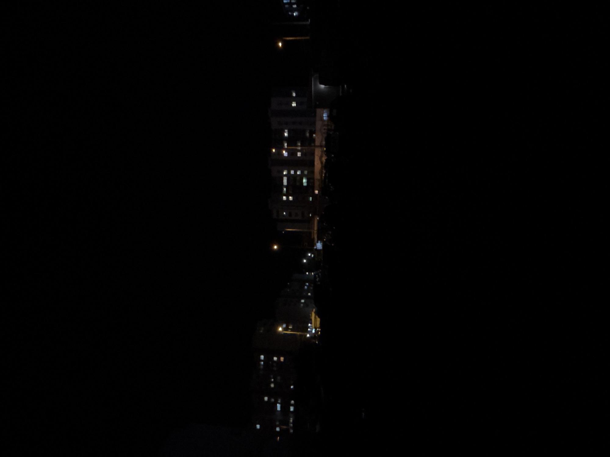 The image depicts a nighttime scene in Burgas, Bulgaria. Buildings line a street illuminated by sporadic streetlights and illuminated windows. The buildings vary in height and architecture, with some appearing to be residential apartments and others possibly commercial structures. No people are visible. The scene appears calm and quiet.