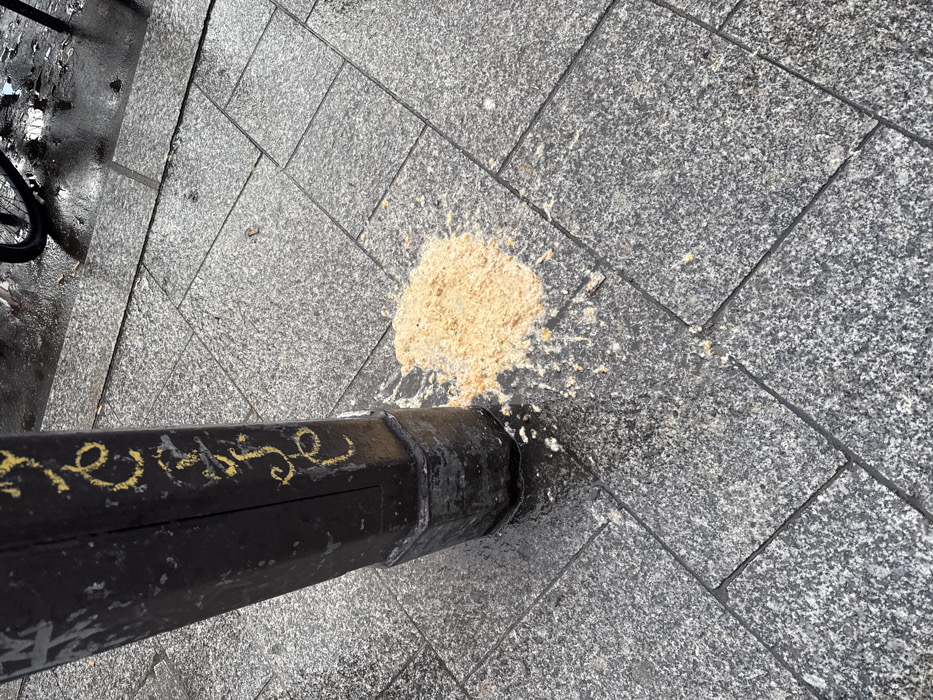 An outdoor urban scene shows a pile of vomit splattered on a grey tiled sidewalk, next to a black pole. The vomit is light beige and granular, spread out from the base of the pole. The pole itself is black, appears metallic or concrete, and has yellow, stylized cursive-like graffiti running along its side. The graffiti is mostly illegible but contains shapes resembling letters such as 'n', 'e', 'r', 'a', 'g', 'e', or 's', possibly hinting at "heritage" or a similar word. The sidewalk is paved with rectangular, mottled grey granite tiles. The ground, particularly on the left side of the image near what appears to be a portion of a bicycle wheel, is wet and reflective, suggesting recent rain or damp conditions. The lighting indicates it is daytime. The overall setting is a public space in a city, specifically Paris, France as per context. No people are visible, and the primary activity suggested is the unfortunate occurrence of someone being sick.