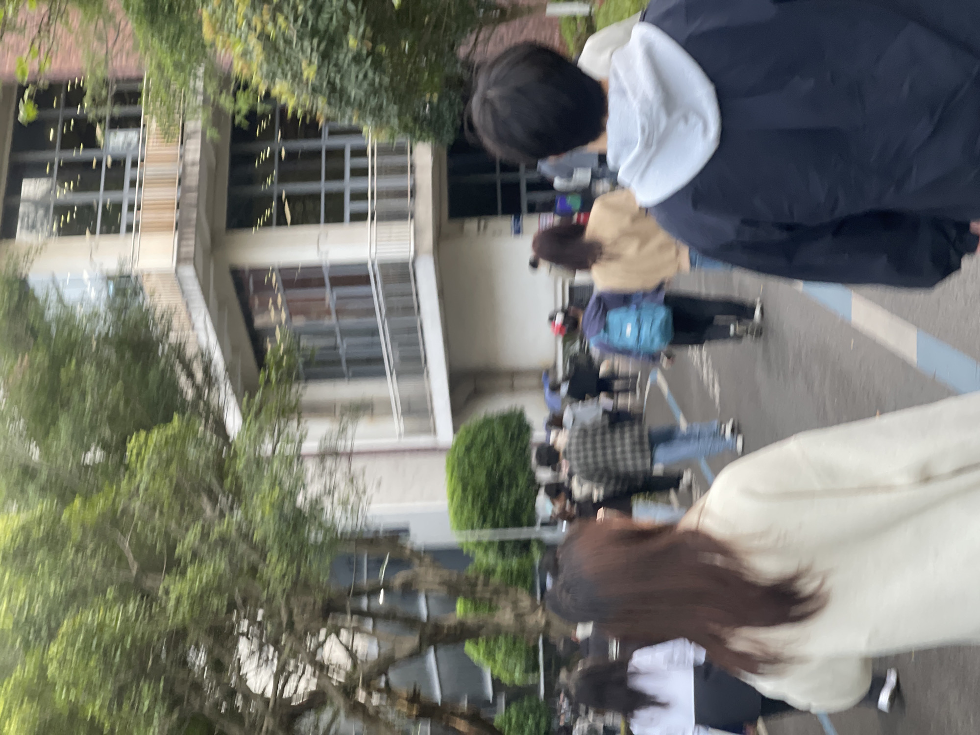 The image depicts a group of people walking along a path in the Chongqing High-tech Zone, China. A multi-story building with many windows is visible on the left side of the image, partially obscured by trees. The path is marked with blue lines. People are walking along the path. They are walking in the same direction.