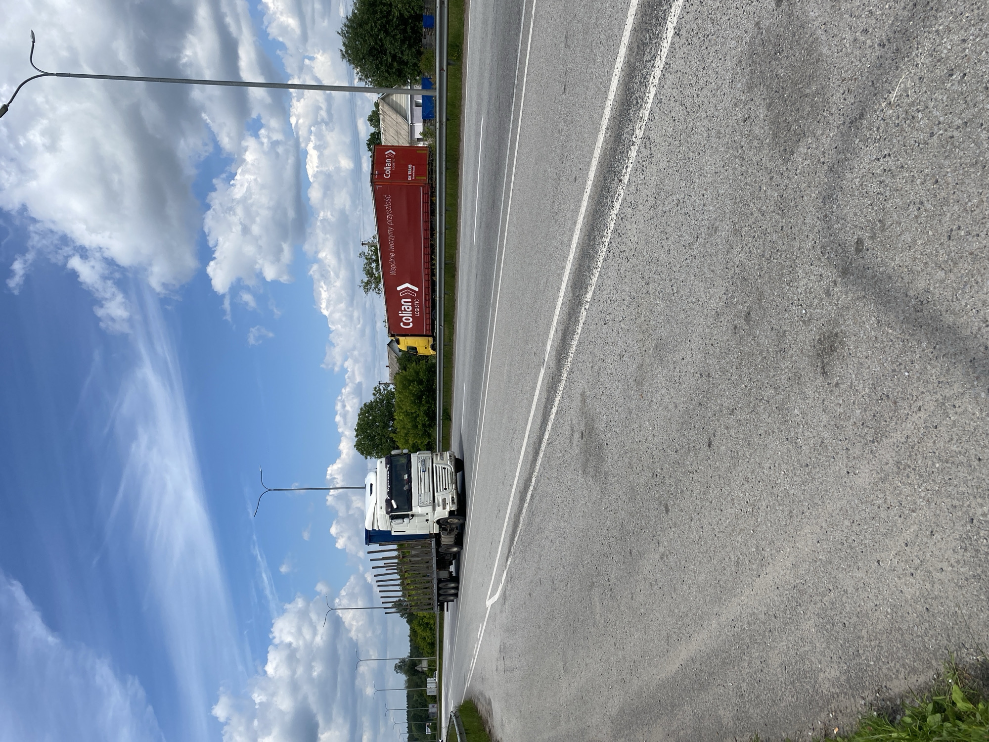 The image shows a roadside scene in Velyki Kosharyshcha, Ukraine. The main subject is a white semi-truck with a long, red trailer. The trailer has "Colian LOGISTIC" written in white text, and the phrase "Wspólne tworzymy przyszłość" (Together we are creating the future) is also visible on its side. The truck is driving on a paved road with white lane markings.
The setting appears to be a highway or a major road passing through a rural or semi-rural area. On the left side of the road, there are trees, some houses, and utility poles with streetlights. The sky is blue with scattered white clouds, suggesting it's daytime, likely late morning or afternoon. The weather appears to be fair and sunny.
The image is oriented vertically, with the road and truck running from bottom to top. The perspective is from a slightly elevated position, looking down at the road. There are no people visible in the image. The overall impression is of a typical day of transportation and logistics on a Ukrainian road.