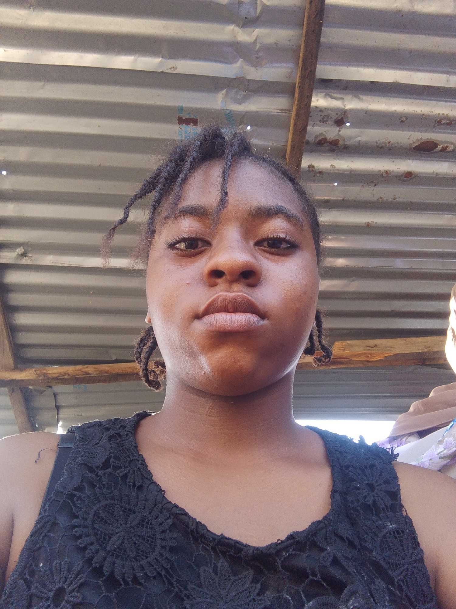 A young woman is pictured in a close-up, slightly low-angle shot. She has dark skin, brown eyes, and dark braided hair. She wears a black lace top. The background consists of corrugated metal and wooden beams, suggesting a simple structure. This could indicate a market area or a modest residential setting in Lokoja, Nigeria.