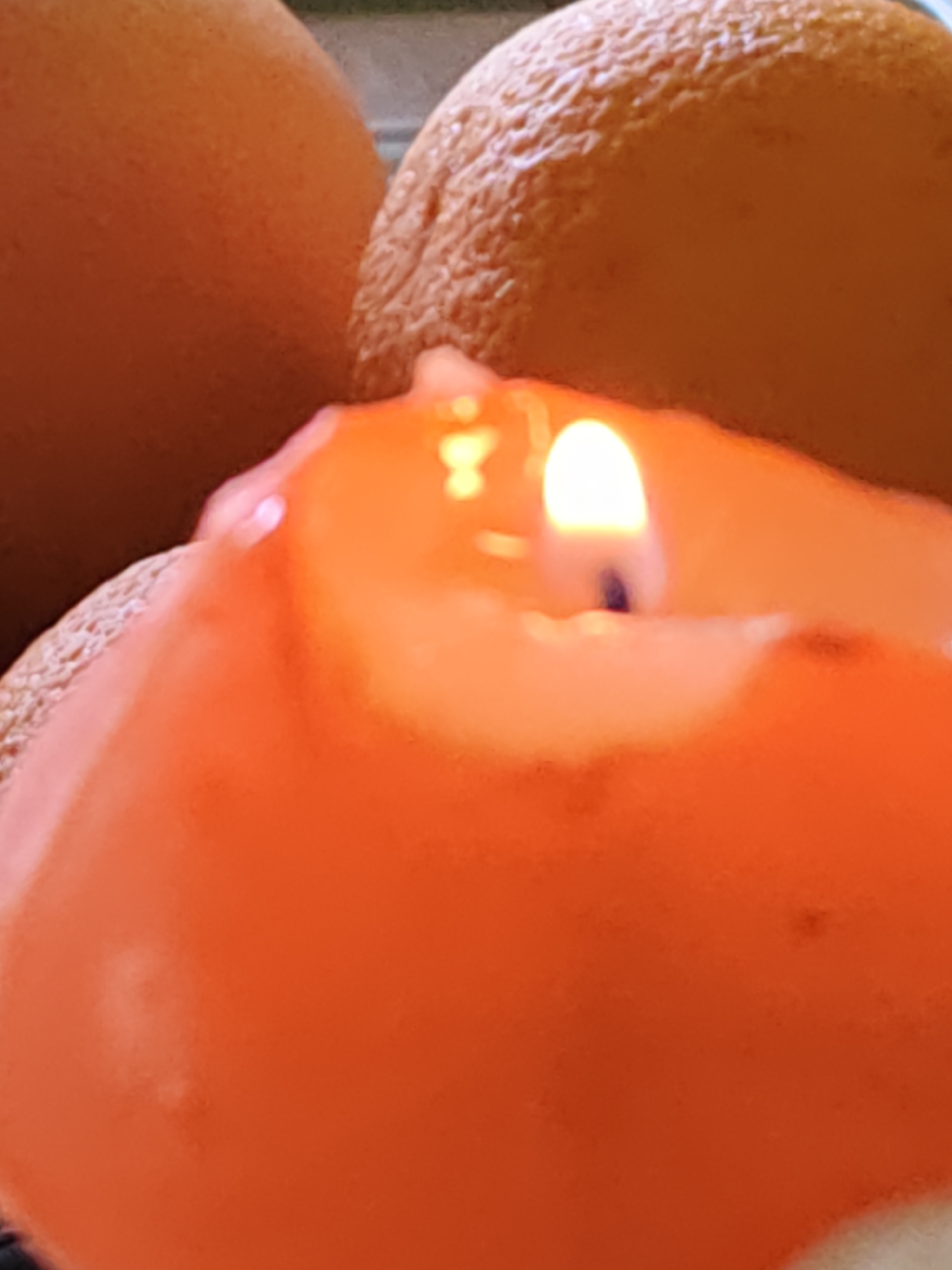 The image shows a close-up of a lit orange candle with a small flame. In the background, out of focus, are the rounded surfaces of what appear to be oranges. The scene is brightly lit, with warm tones dominating the color palette. There is no indication of the city of Rostov-on-Don, Russia, or any other specific location.
