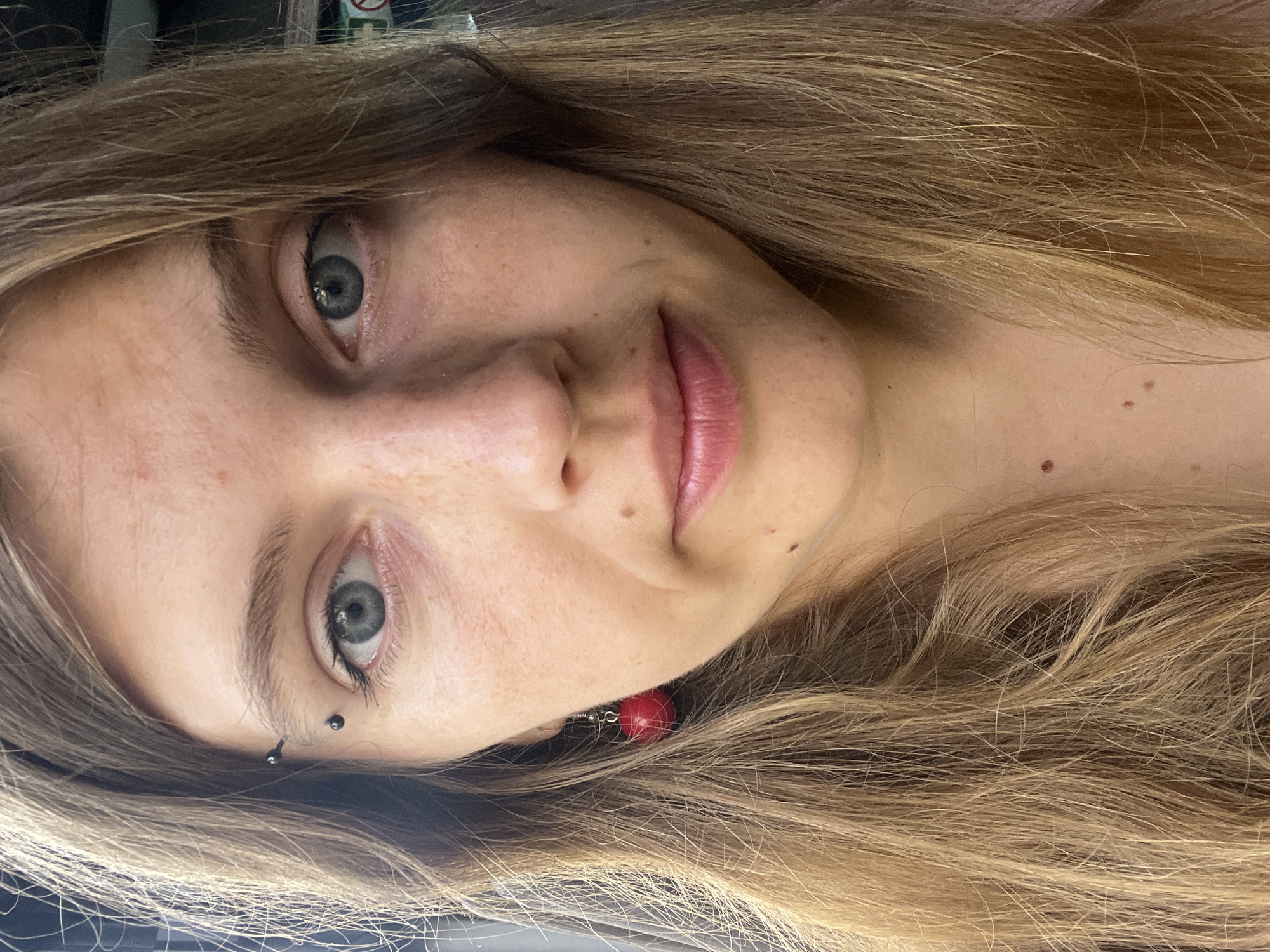 A close-up, head-on, eye-level photograph shows a young Caucasian woman with long, light brown hair with blonde highlights. She is wearing a black industrial piercing in her left eyebrow and a red, spherical earring in her left ear. Her skin is fair and appears to have freckles. She has light blue eyes and thin, rosy lips. The background is blurred, but appears to be an indoor setting with some signage visible on the right side. The image is well-lit, likely by natural light. The location is not specified in the image or its surrounding context.